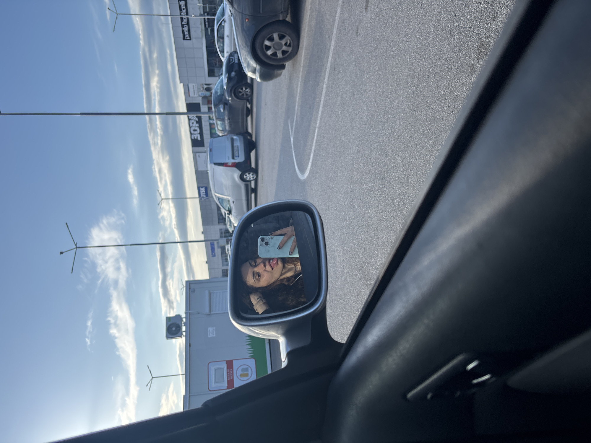 The image depicts a parking lot scene in Sofia, Bulgaria, as indicated by the Cyrillic text "3OPA" on a building in the background. The viewpoint is from inside a car, focusing on the side mirror. The reflection in the mirror shows a young woman holding a smartphone, presumably taking a selfie. Outside the car, several other vehicles are parked in designated spaces. A portable office or storage container is visible near the car, with some signage attached. The sky is blue with scattered clouds, suggesting a sunny day. The pavement is asphalt and marked with white parking lines. Tall streetlights with multiple arms are also visible in the background.