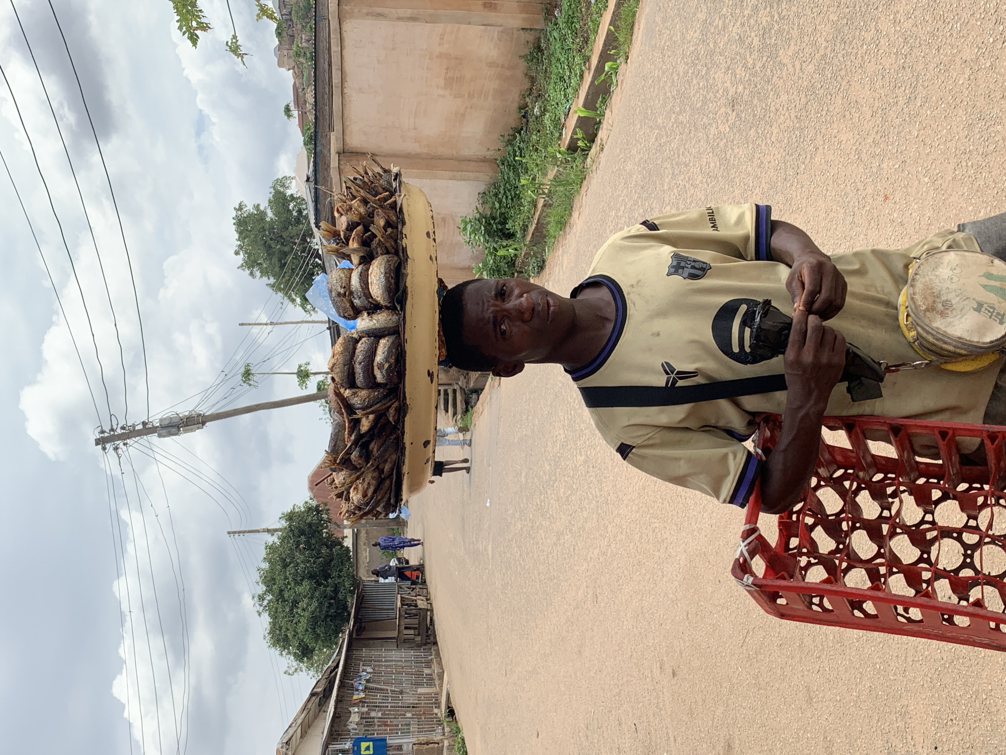 This image captures a street scene in Jos, Nigeria, under an overcast sky. The central figure is a young Black male, positioned in the foreground, facing the camera. He wears a light tan short-sleeved t-shirt with dark blue trim on the collar and sleeves, featuring a dark "Y" shaped logo on the left chest, a shield emblem on the right chest, and "AMBILI" text on the right sleeve. He has a dark shoulder strap across his chest and a small, worn, circular yellow and white bag with green graphics hanging from his left hip. His hands are clasped, holding a small dark object. Balanced on his head is a large, shallow, round, yellow-tan metal tray filled with various smoked fish, some stacked and others loose, along with a light blue plastic bag containing unspecified items. To the left of the man, partially visible, is a red plastic crate with an open grid pattern, secured at the top with white tape.
The scene unfolds on an unpaved, light brown road that extends into the distance. Along the left side of the road, a row of simple, single-story buildings with corrugated metal roofs and walls is visible; clothes hang on a line in front of one structure, and a blue square sign with a yellow arrow is attached nearby. Further down on the left, a large, dense green tree stands. On the right side of the road, a plain, light brown wall runs parallel to the street, with sparse green vegetation growing in a narrow ditch area in front of it. In the background, rocky hills with some structures and vegetation are discernible under the cloudy sky. Multiple wooden utility poles with numerous power lines crisscross the upper portion of the frame. Two male figures walk away from the camera further down the road, and two sets of legs are visible beneath the tray on the right side of the road.