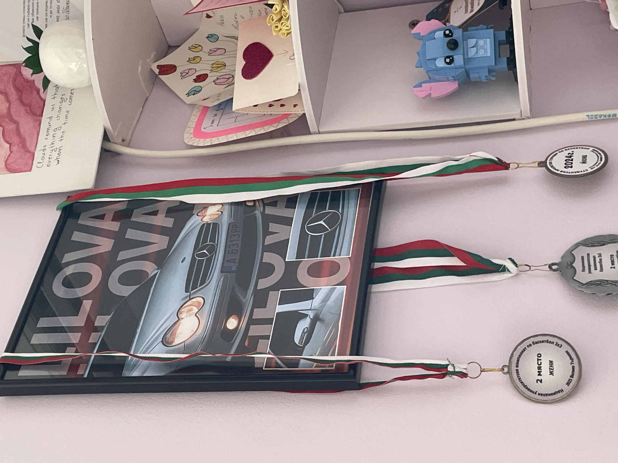 The image shows a shelf with various items, including children's drawings and a LEGO figure, with a framed picture hanging below. The framed picture, displaying an image of a Mercedes-Benz car, is suspended by three medals, each with ribbons featuring the Bulgarian flag colors (white, green, and red). The medals indicate accomplishments in basketball, specifically a second place finish in a women's tournament at the National University level. One medal mentions "Veliko Tarnovo 2023", identifying the location as the city of Veliko Tarnovo, Bulgaria. Another medal shows "Veliko Tarnovo 2024", with the inscription in Cyrillic.