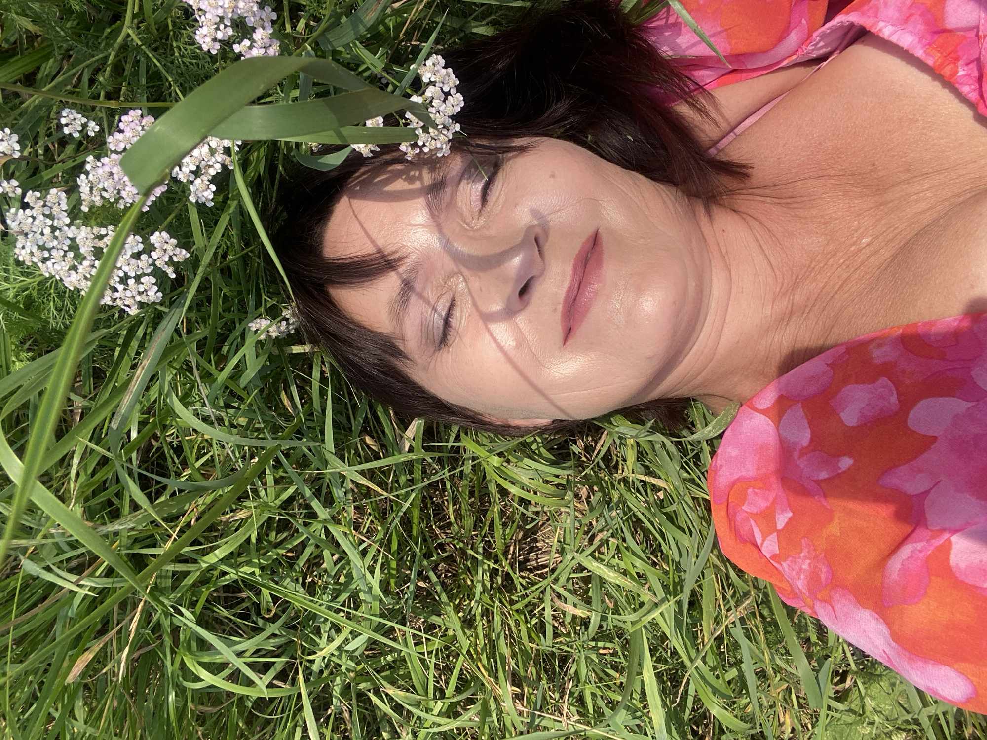 A woman is lying in the grass with her eyes closed, her face tilted towards the sun. She is wearing a bright pink and orange floral dress. Small white flowers are interspersed with blades of green grass around her head. The lighting suggests it is a sunny day. The scene is outdoors and appears to be a natural, grassy area. There is no indication of the city of Novokuznetsk, Russia.
