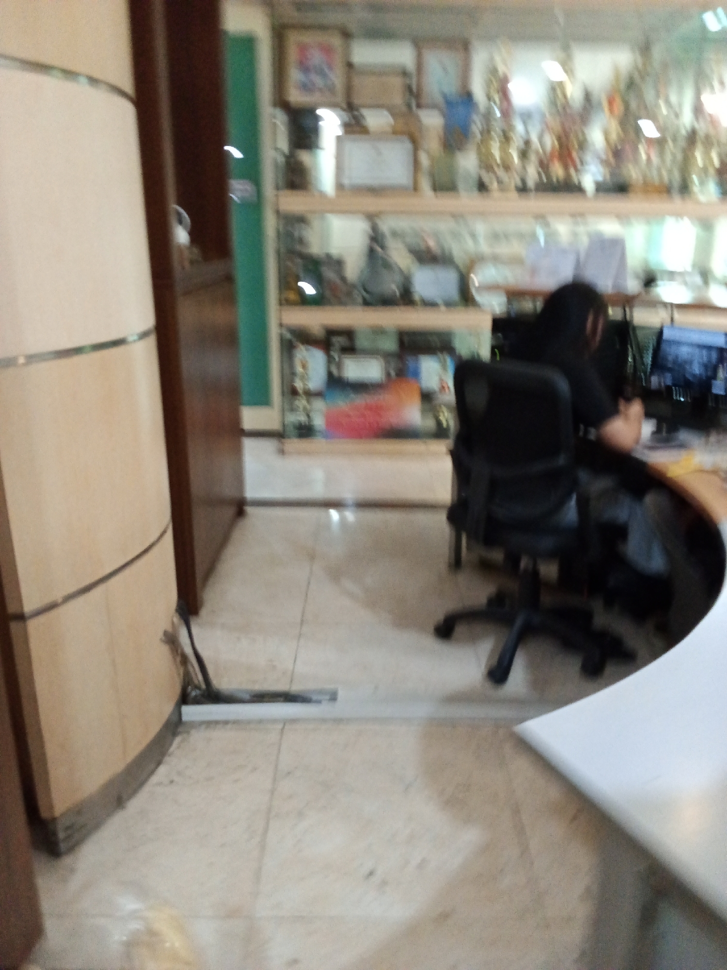 In an office environment, a person with dark hair is seated at a desk with a computer monitor. They are wearing dark clothing. A black office chair is positioned in front of the desk. Shelving units filled with trophies and various items are visible in the background. The floor is tiled. The scene appears to be in Bandung City, Indonesia, based on the context provided.
