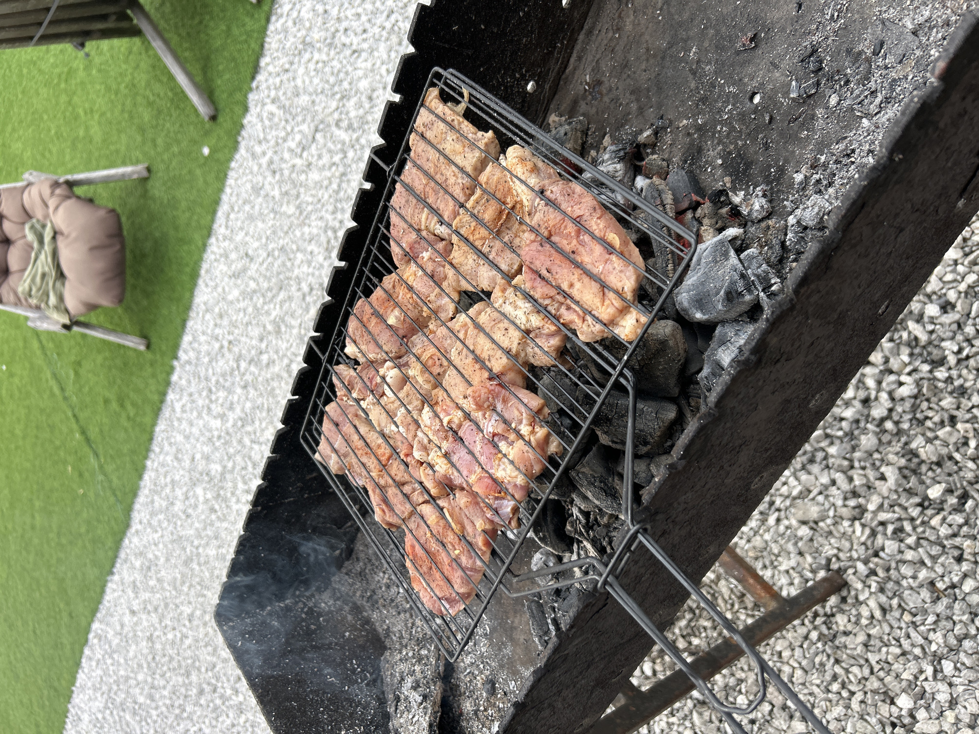 In Sheregesh, Russia, a barbecue grill is loaded with rows of raw, seasoned meat slices arranged on a metal rack over hot coals. The grill is situated on a gravel surface, with a patch of artificial turf visible in the background. To the left of the grill, a small amount of smoke rises. In the background, a lounge chair with a cushion and a folded green fabric is placed on the artificial turf.
