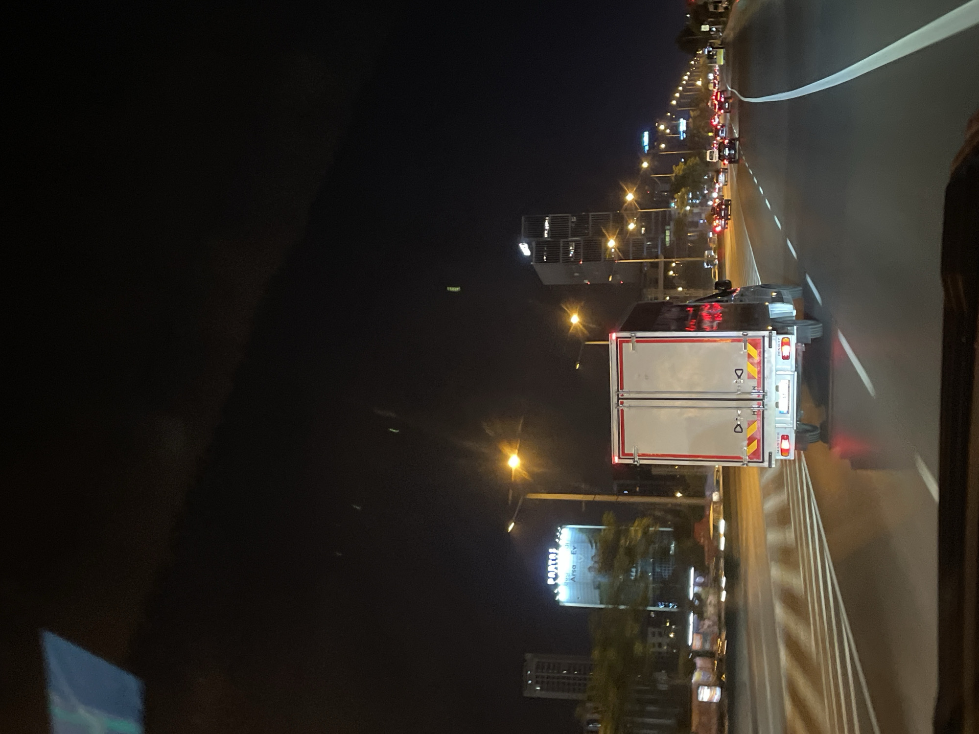 The image captures a night scene on a multi-lane road in Ataşehir, Turkey. A white box truck with red and yellow reflective tape on its rear is prominently featured in the foreground, traveling in the right lane. The truck has the brand "RENAULT" visible on its rear bumper. On the left side of the road, several buildings are illuminated, including a skyscraper with a sign that appears to read "PARTOS" at the top. Adjacent to it is another tall building. The road surface is wet, reflecting the lights. Traffic lights and streetlights line the road, illuminating the night sky. Other vehicles, including cars, are visible in the distance, moving in the same direction. The overall environment suggests an urban setting at night.