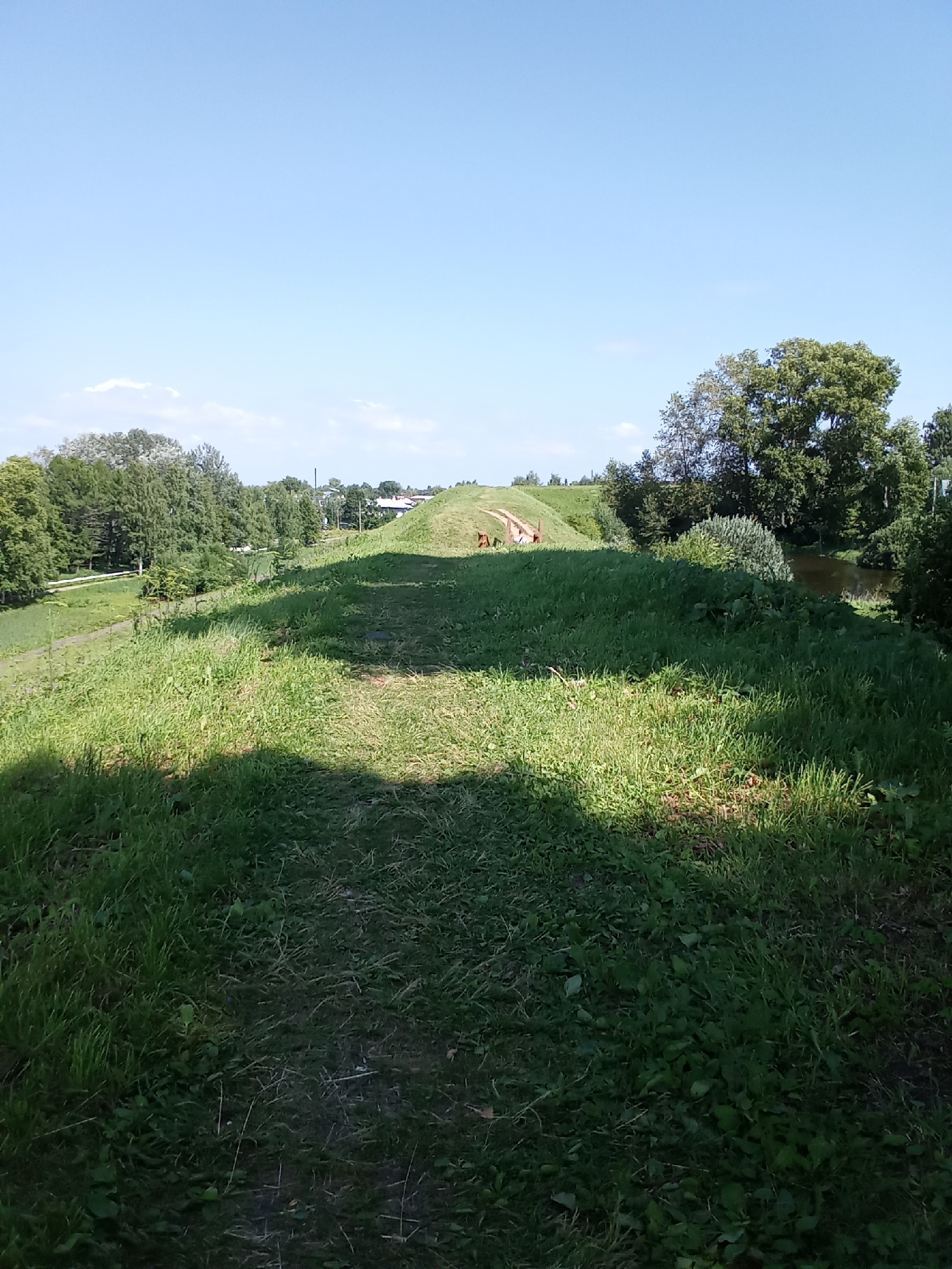 An eye-level, medium-shot photograph captures a sunlit, grassy embankment with a narrow, dirt path leading upwards and to the left. The path, in the foreground, is partially in shadow, with tall grass on either side. The embankment itself is covered in lush green grass, with scattered patches of sunlight and shadow. In the middle ground, the path curves to the left and appears to lead towards a structure or bridge made of weathered, orange-brown material. Two small, indistinct figures, possibly people or animals, are visible on the path near this structure. Beyond the embankment, a dense line of green trees stretches across the horizon, with a few buildings and power lines interspersed. To the right of the embankment, a dark, still body of water, likely a river or canal, is visible, bordered by trees. The sky above is a clear, bright blue with a few wispy white clouds. The overall impression is of a natural landscape on a sunny day in a park or natural area within the city of Belozersk, Russia.