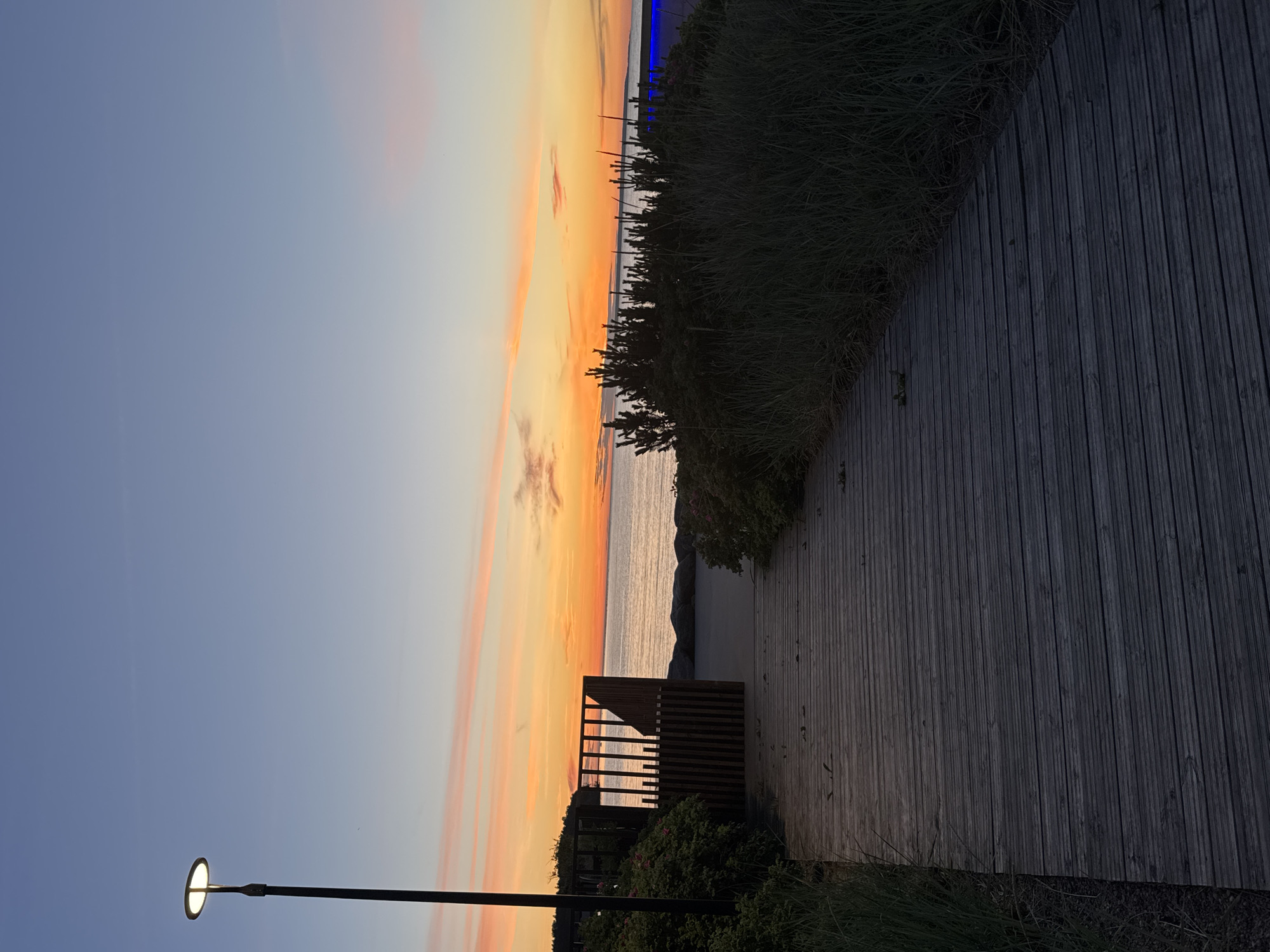 A wooden boardwalk leads towards a beach at sunset. The sky is a gradient of blue and orange, with streaks of pink clouds. A modern lamp post stands to the left of the boardwalk. To the right, tall grass and bushes line the path. In the distance, the calm sea stretches to the horizon, bordered by a line of rocks. A wooden structure with vertical slats is visible near the start of the beach access. The image captures a serene evening scene, likely in a coastal area of Tallinn, Estonia, given the style of architecture and landscaping.
