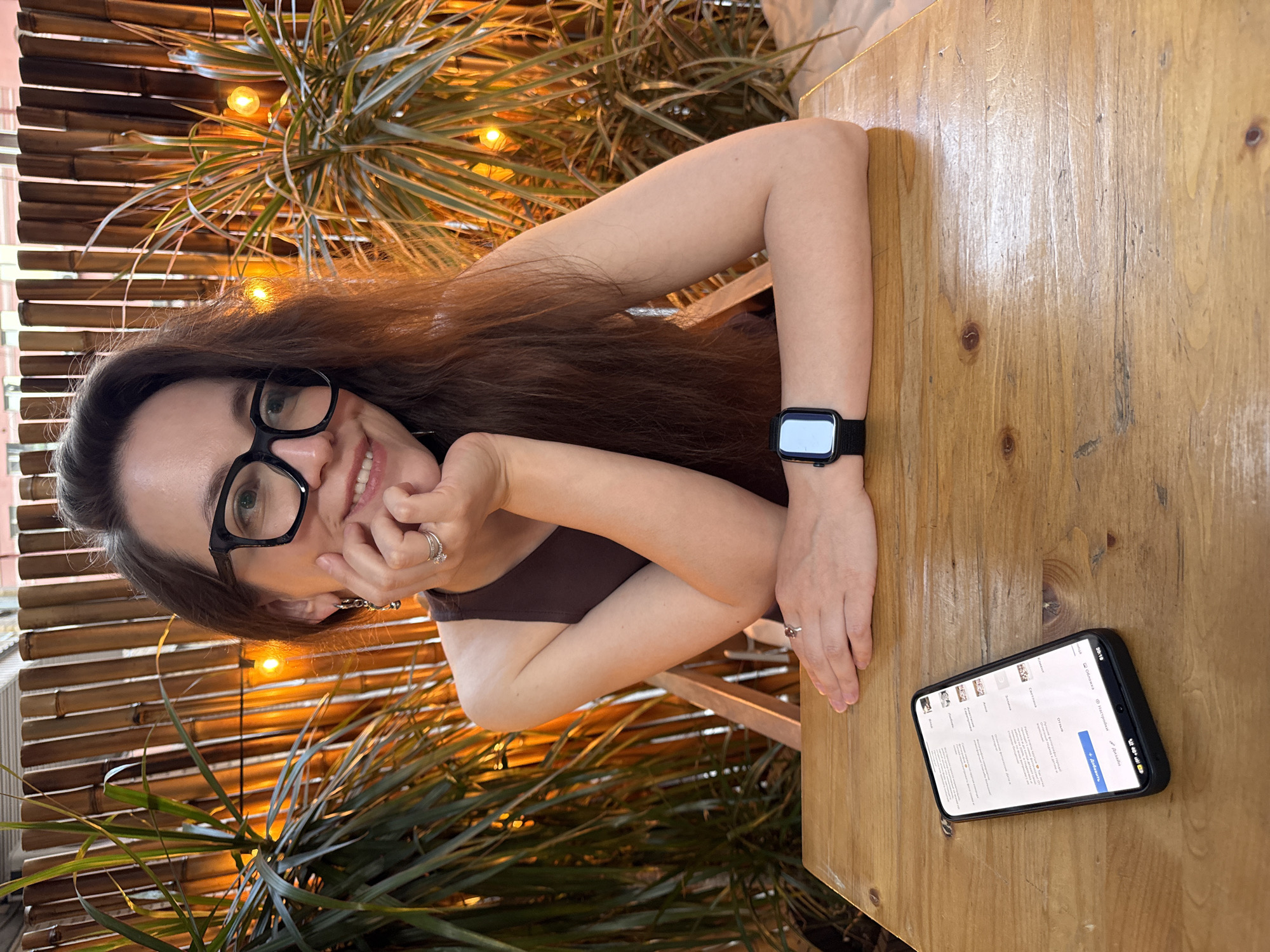 A woman with long brown hair and black-framed glasses is seated at a wooden table. She has a black smartwatch on her left wrist and rings on her fingers. Her right hand is resting on her chin. A smartphone is lying on the table near her right hand, displaying what appears to be a webpage in Russian. Behind her is a bamboo fence decorated with string lights and a plant. Based on the phone's language settings and the overall aesthetic, the location appears to be a restaurant in Moscow, Russia.