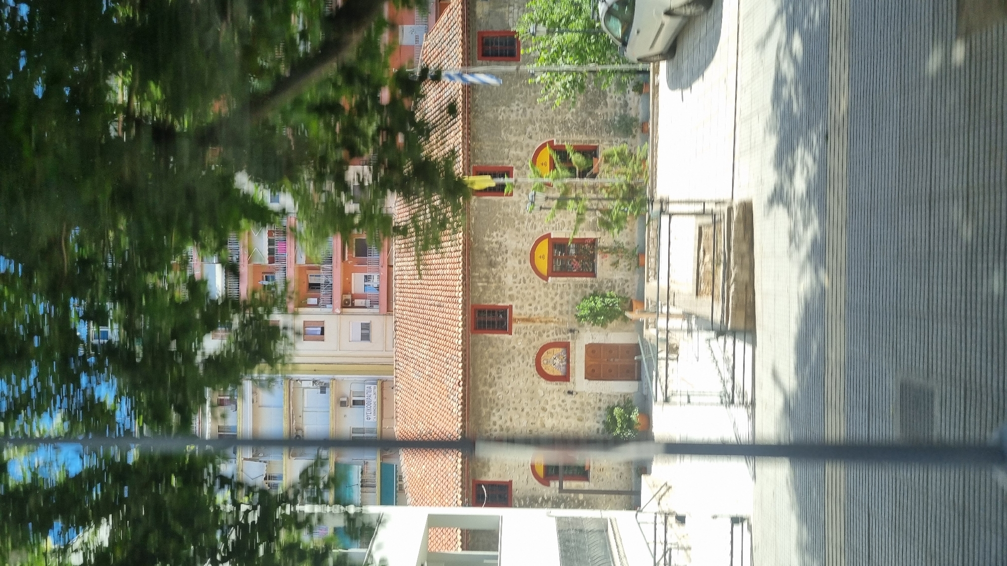 This is a photograph taken from a moving vehicle, likely a bus, looking out at a street scene in Thessaloniki, Greece. The foreground is dominated by blurred green foliage from trees and a vertical dark pole. In the midground, a stone church with a red tile roof is visible. The church has arched windows with red frames and a decorated arched entrance. To the left of the church is a white modern building. Behind the church and modern building, a taller, multi-story apartment building with balconies in various colors (pink, white, and yellow) is visible. A white car is parked on the right side of the image. The ground in front of the church is paved, and there are shadows from trees cast on the ground. The overall impression is a sunny day with movement blur due to the vehicle's motion.