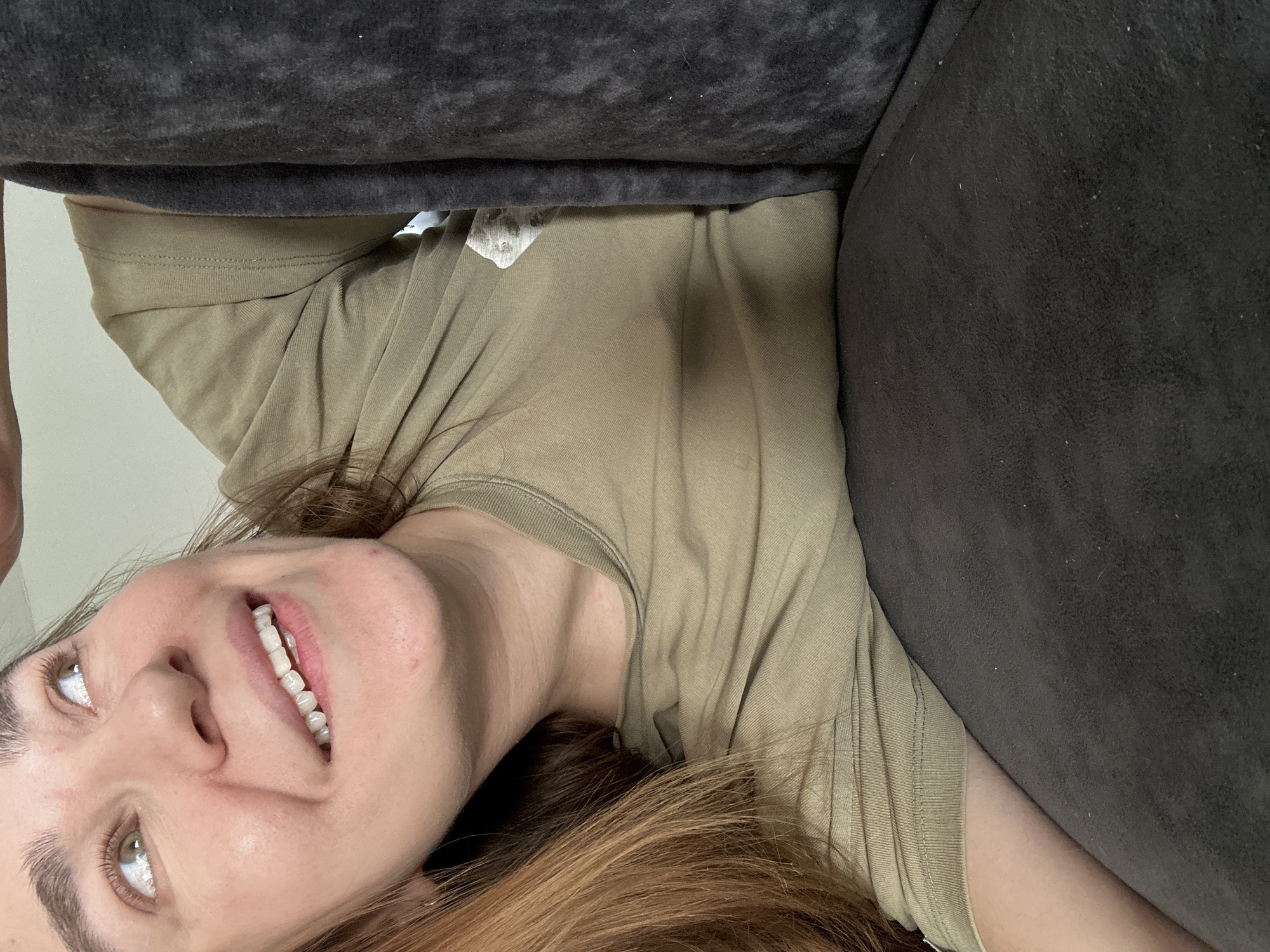 A portrait-style image, taken from above, shows a woman lying on her back on dark-colored upholstered furniture, likely a couch or bed, in Kharnavy, Russia. Her head is positioned towards the left side of the frame, and she is looking upwards, smiling warmly towards the camera. Her light brown hair is spread out around her head and shoulders. She has light-colored eyes and a wide smile, revealing a full set of white teeth. She is wearing a light olive green, short-sleeved t-shirt with a ribbed collar. A subtle, lighter-colored graphic or emblem is visible on the upper chest area of her shirt, and a white label or tag can be seen peeking out from the inside collar. The dark, textured fabric of the furniture, which appears to have some lint or dust on its surface, frames much of the shot, with a light-colored wall or surface visible in the top left corner. The woman appears relaxed and happy, conveying a sense of ease.