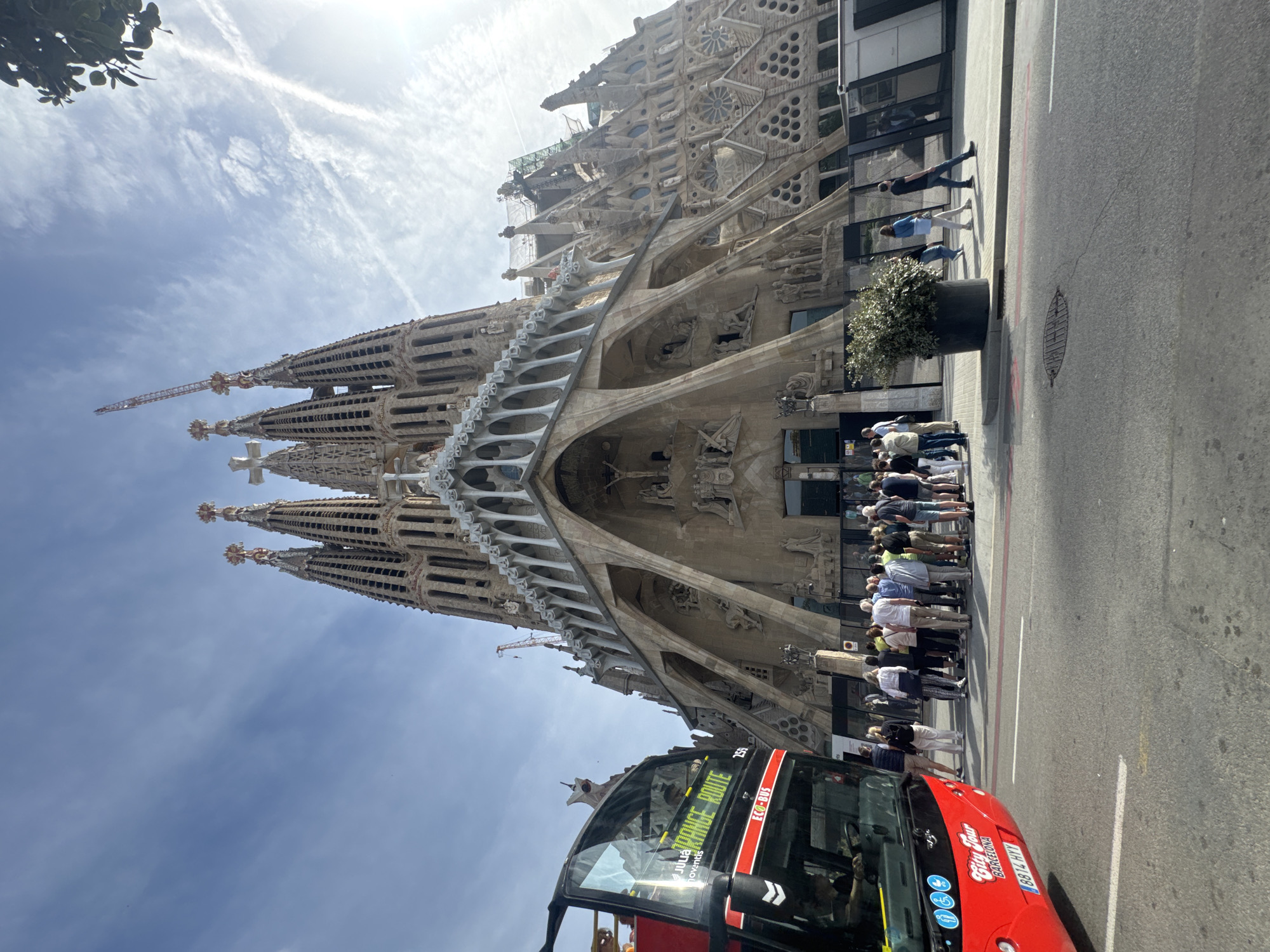 This image captures a vibrant street scene in Barcelona, Spain, dominated by the iconic Sagrada Familia cathedral. The immense, intricately detailed basilica stands as the central subject, showcasing its unique Gothic and Art Nouveau architectural style with numerous spires, sculptures, and facade elements. Construction is ongoing, evidenced by a visible crane near one of the towers.
In the foreground, a bright red open-top double-decker tour bus is prominently featured, heading towards the viewer. Visible text on the bus includes "City Tour Barcelona," "Julia Travel," "ECO-BUS," and "ADVANCE ROUTE." The license plate "8814 HVY" is also visible.
Along the sidewalk, a substantial queue of people is lined up, presumably waiting to enter the Sagrada Familia or for the bus. They are dressed in casual attire suitable for warm weather. A large potted plant sits between the queue and the street. Beyond the people, a modern building with large glass windows reflects the surroundings.
The weather appears bright and sunny, indicative of daytime. The sky is a clear blue with some scattered white clouds and distinct contrails from an aircraft, adding to the bright atmosphere. The overall scene depicts a bustling tourist area, with visitors drawn to one of Barcelona's most famous landmarks.