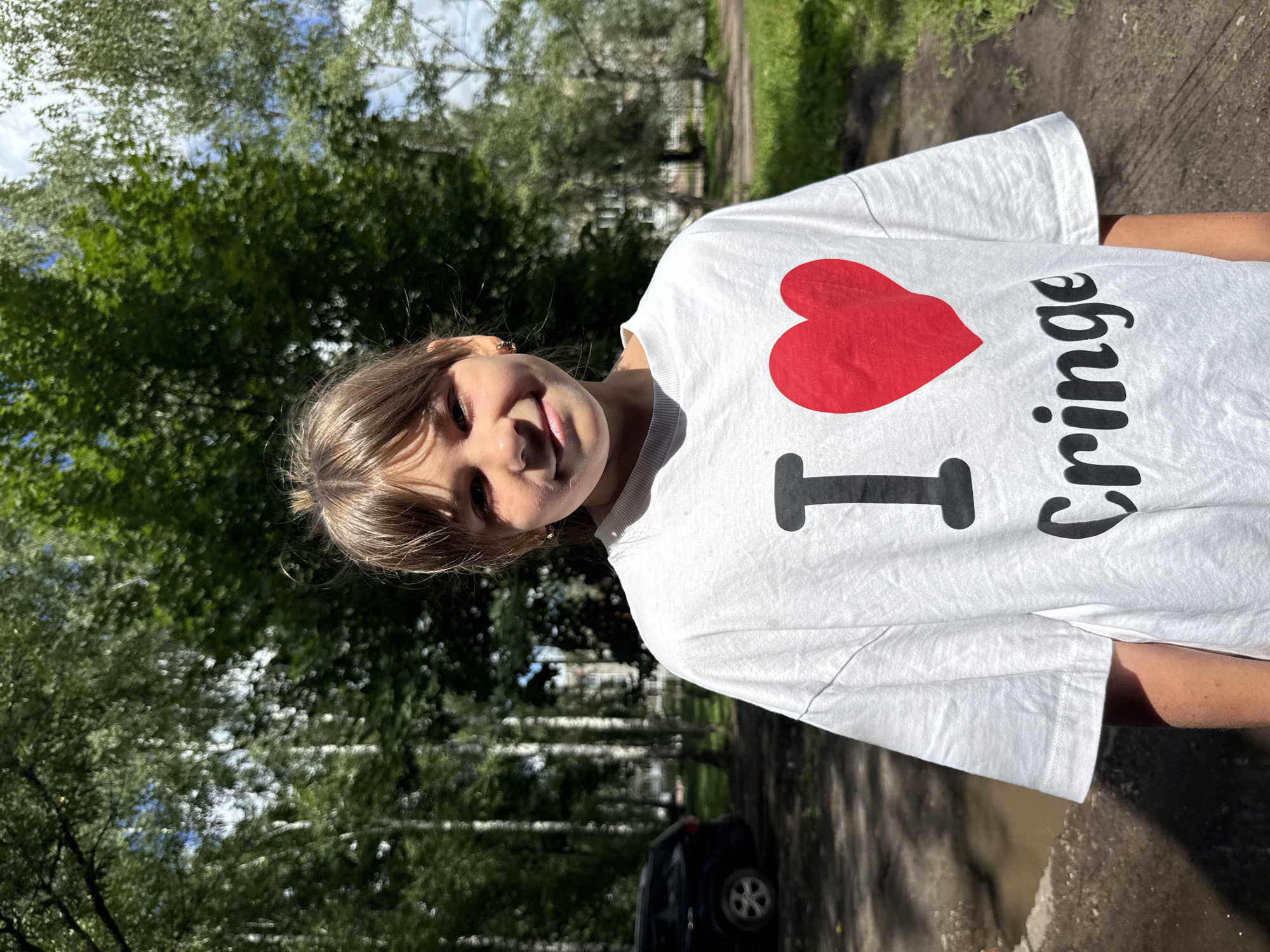A young girl stands outdoors in front of a line of trees and a black SUV. She is wearing a white t-shirt with a large red heart and the word "Cringe" printed on it. The girl has light brown hair pulled back in a messy bun and is smiling at the camera. The background is a mix of trees and sky, suggesting a sunny day. The scene is located in the city of Rusanovo, Russia.