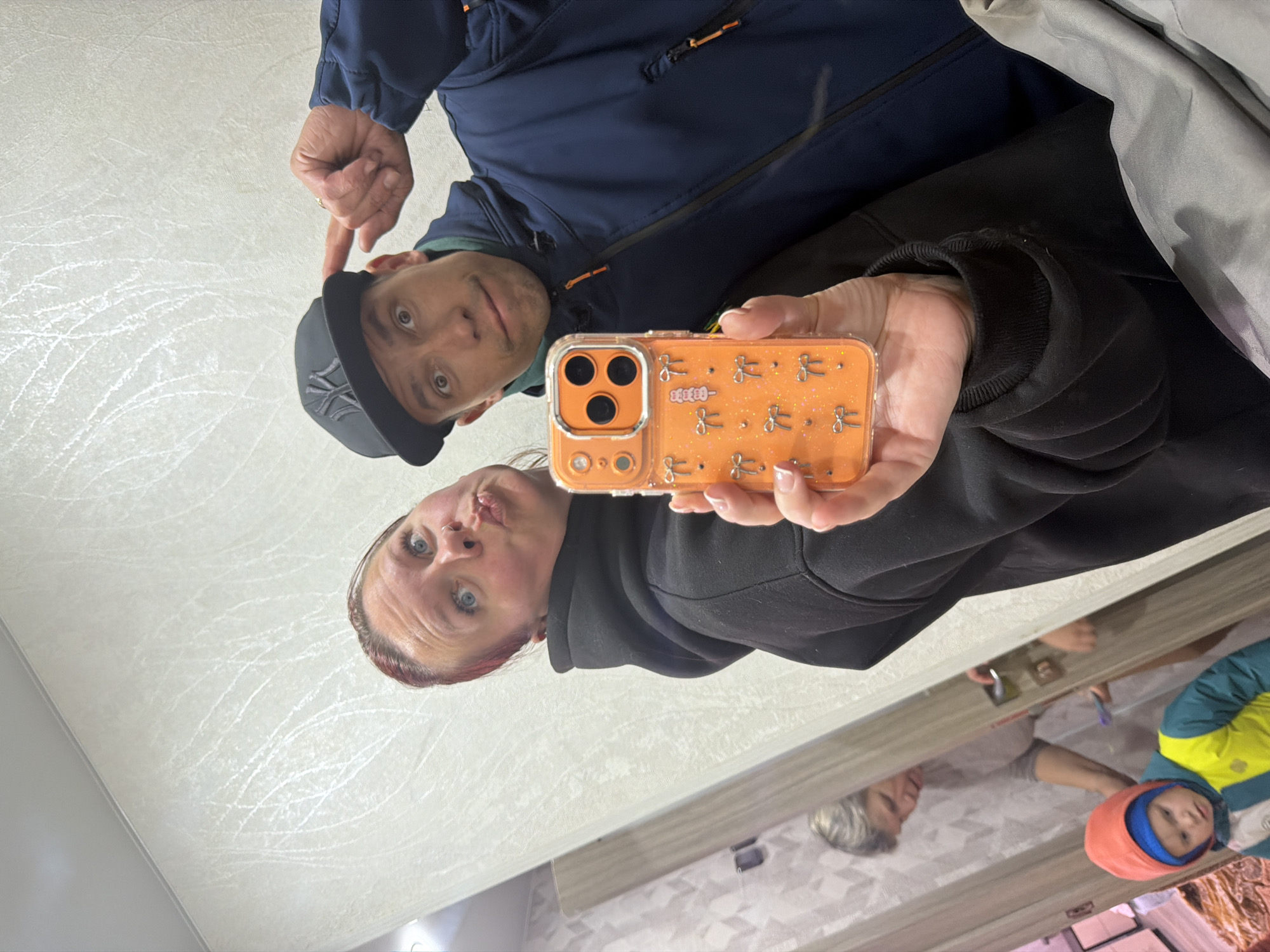 A selfie taken in a mirror shows two adults. One adult is wearing a dark-colored hoodie and is holding an orange cell phone with a clear case decorated with bow ties. The other adult is wearing a blue jacket and a dark baseball cap with a logo. In the mirror's reflection, a child wearing a brightly colored jacket and hat and another adult with gray hair can be seen. The location is unclear, but it is possibly in Chaikovsky, Russia.