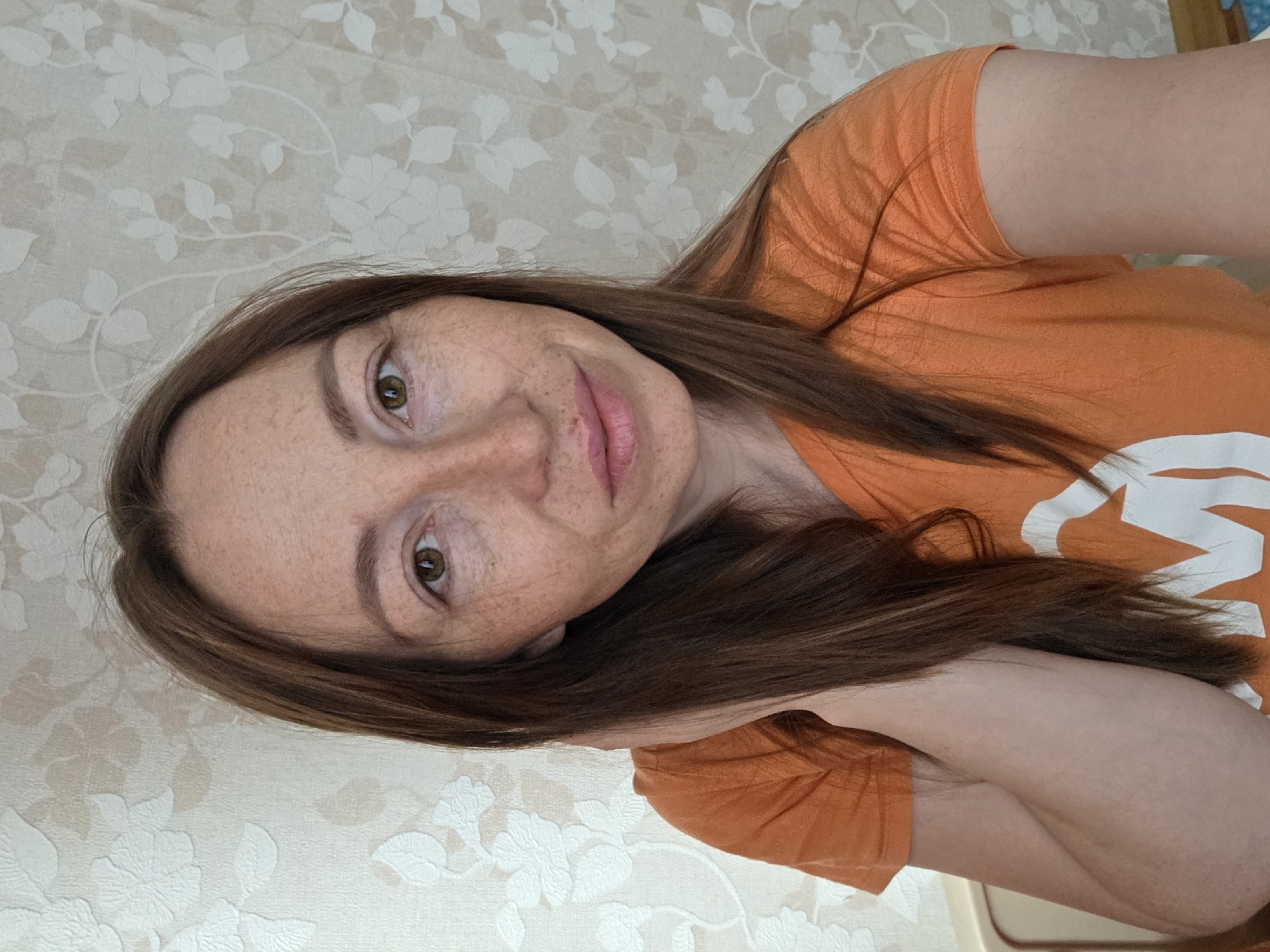 This close-up selfie, taken in Chaikovsky, Russia, features a person, likely a woman, with fair skin and numerous freckles across her face. She has long, straight, brown hair that cascades over her shoulders and down her arm. Her eyes are brown, and her lips have a soft pink hue, possibly from lip gloss. She is wearing a plain orange t-shirt with a large, partially visible white graphic or stylized letter on the front.
The person is looking directly at the camera with a calm, relaxed expression and a slight, gentle smile. The image is rotated 90 degrees counter-clockwise, suggesting the photo was taken while lying down or with the camera held at an unconventional angle.
The background consists of light-colored wallpaper featuring a subtle, embossed or textured floral pattern of white or very light beige flowers and leaves against a cream or light beige background. The setting appears to be indoors. There are no clear cues for the time of day or weather. The visible white graphic on the orange t-shirt is a distinct, large, stylized letter, though its full form is not discernible.