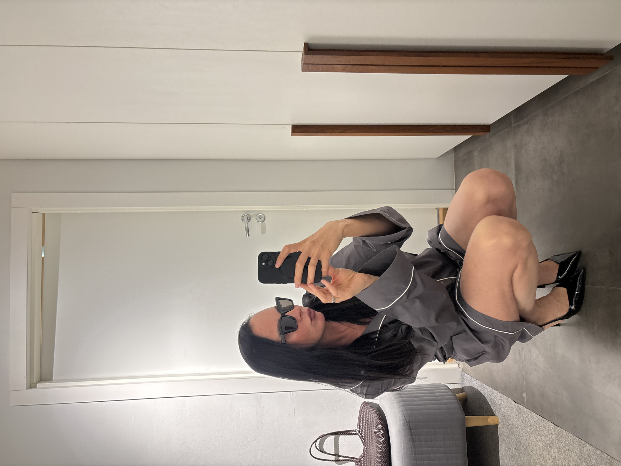 A woman is taking a selfie in what appears to be a dressing room or walk-in closet. She is kneeling with her legs to the right side of the frame. She wears black high-heeled shoes, a gray robe with white piping, and dark sunglasses. Her dark hair is long and partially obscures her face. She is holding a black smartphone with both hands, presumably to take the photo. Behind her, a gray quilted ottoman and a woven handbag are visible. The room has a white door and white walls, with horizontal wooden trim near the top. The floor is gray. It is not possible to confirm the location within Chaikovsky, Russia, based solely on this image.