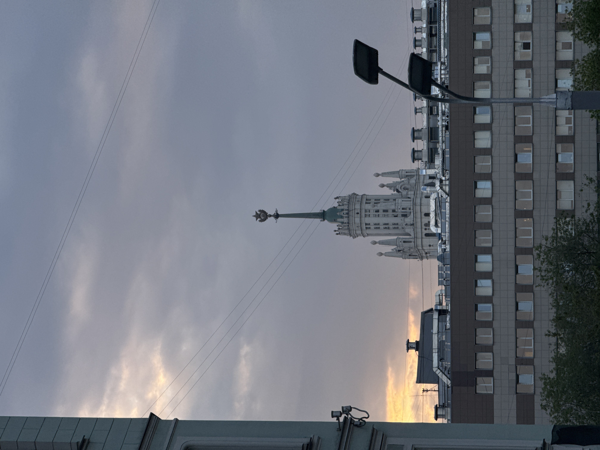 The image shows a cityscape in Moscow, Russia, at twilight. Dominating the skyline is one of the Seven Sisters skyscrapers, with its distinctive Stalinist architecture, including a spire topped with a star. Adjacent to the skyscraper is a more modern, multi-story building with a grid of windows. A street light stands between the viewer and the buildings. Overhead, power lines crisscross the sky against a backdrop of clouds. The lower portion of the image includes a glimpse of the building from which the shot was taken, showing a window frame and part of the roof. The overall atmosphere is muted, with the golden hue of sunset peeking through the clouds.