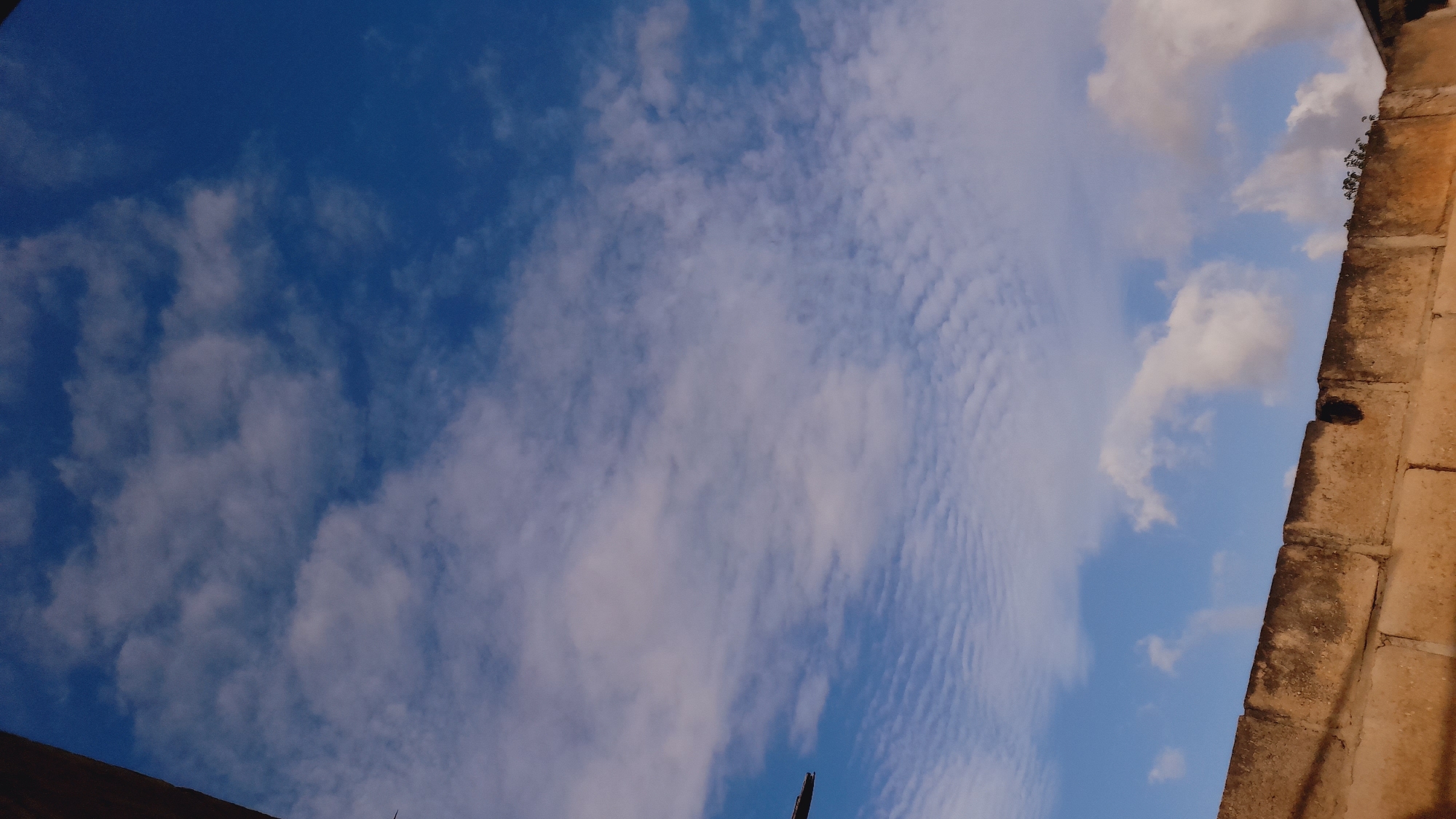 The image depicts a section of a brick wall against a bright blue sky filled with wispy white clouds. The wall appears aged and weathered, constructed from roughly hewn rectangular blocks. The sky is predominantly a deep blue, with scattered altocumulus and cirrocumulus clouds forming delicate patterns. The lighting suggests it is daytime, likely late morning or early afternoon, with sunlight illuminating the scene from above. No people or specific landmarks are visible, however, the style of the brickwork and the clear sky are consistent with the environment found in coastal areas of Venezuela, such as Juan Griego, known for its historical significance and maritime atmosphere.