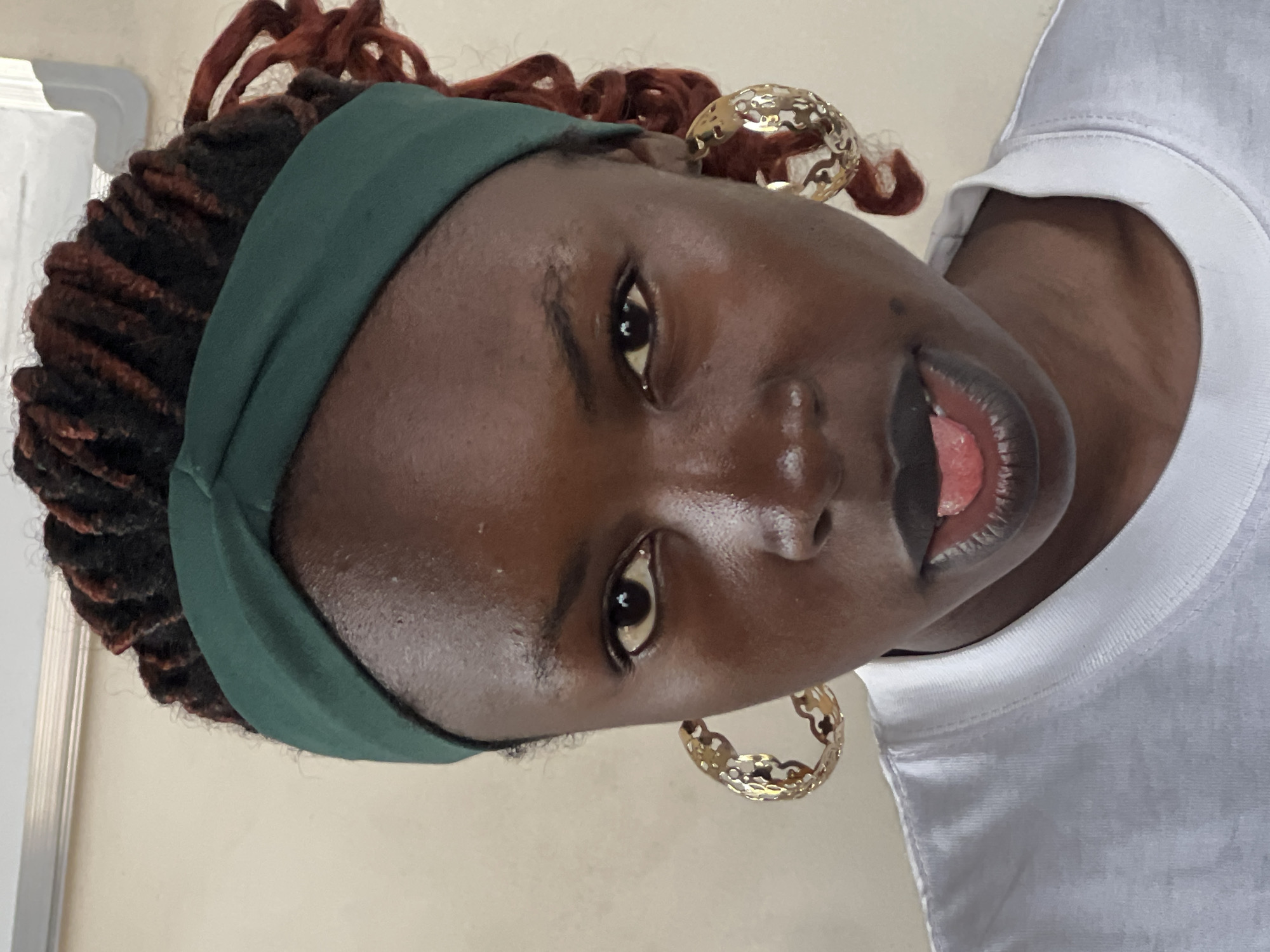 A close-up, head-on, waist-up shot captures a Black woman with dark skin and auburn-tipped dreadlocks, pulled back by a dark green fabric headband. She is wearing large, ornate gold hoop earrings and a white t-shirt. Her lips are dark, and her tongue is slightly extended, touching her lower lip. The background is a plain, light beige wall. The lighting is natural and appears to be coming from the front, creating some shine on her face. No specific geographical markers for Jos, Nigeria, are visible in the image.