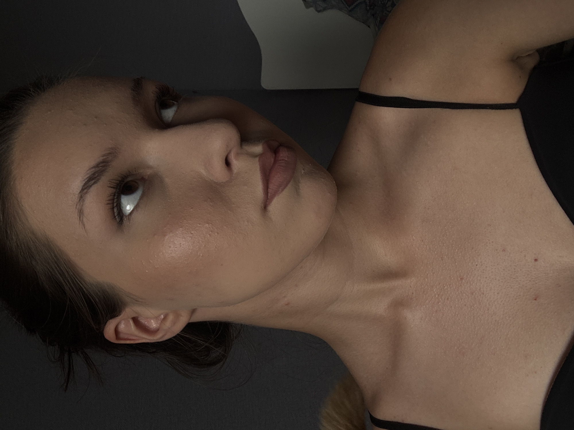 The image shows a close-up of a young woman's face and upper body. She has fair skin, brown eyes, and brown hair pulled back. She is wearing a black camisole. The background is dark and indistinct. There are no discernible elements in the image that would suggest a location in Vishnevka, Russia.