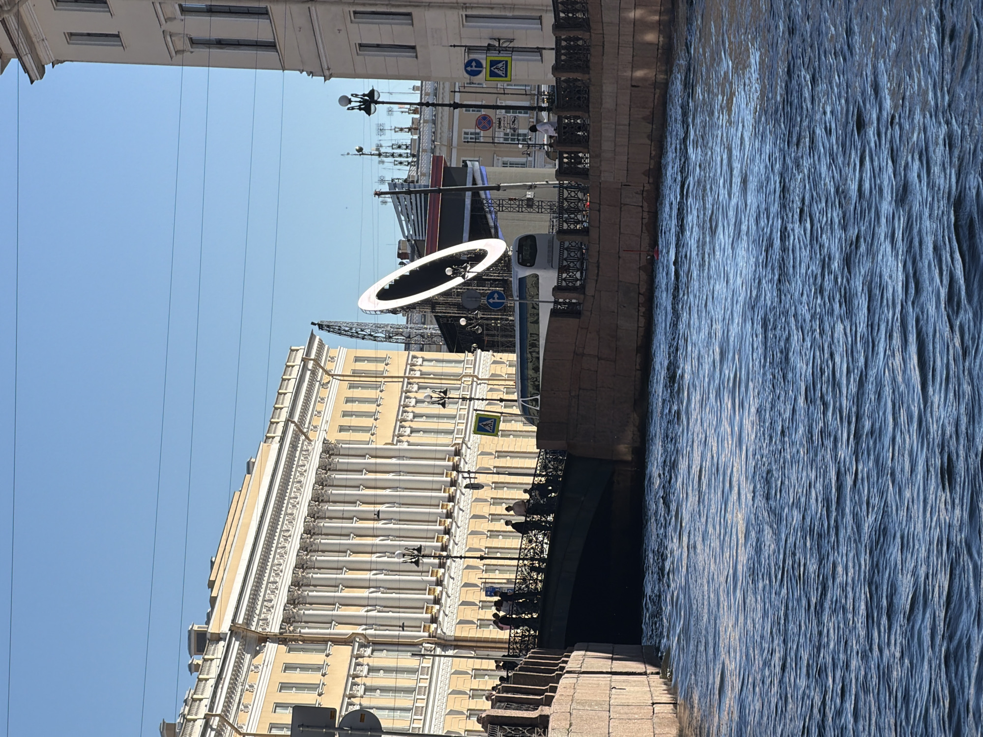 This is a photograph of a canal with a bridge spanning it in Saint Petersburg, Russia. The canal is filled with dark blue, rippling water. The bridge, made of stone and ornate metal railings, is crowded with people. On the far bank, there is a large, ornate yellow building with classical columns and many windows. Behind the building, a large, illuminated ring-shaped structure is visible, along with scaffolding and what appears to be a stage set. Traffic lights and street lamps are also present. The sky is clear and blue. The scene appears to be a public area with architectural significance.