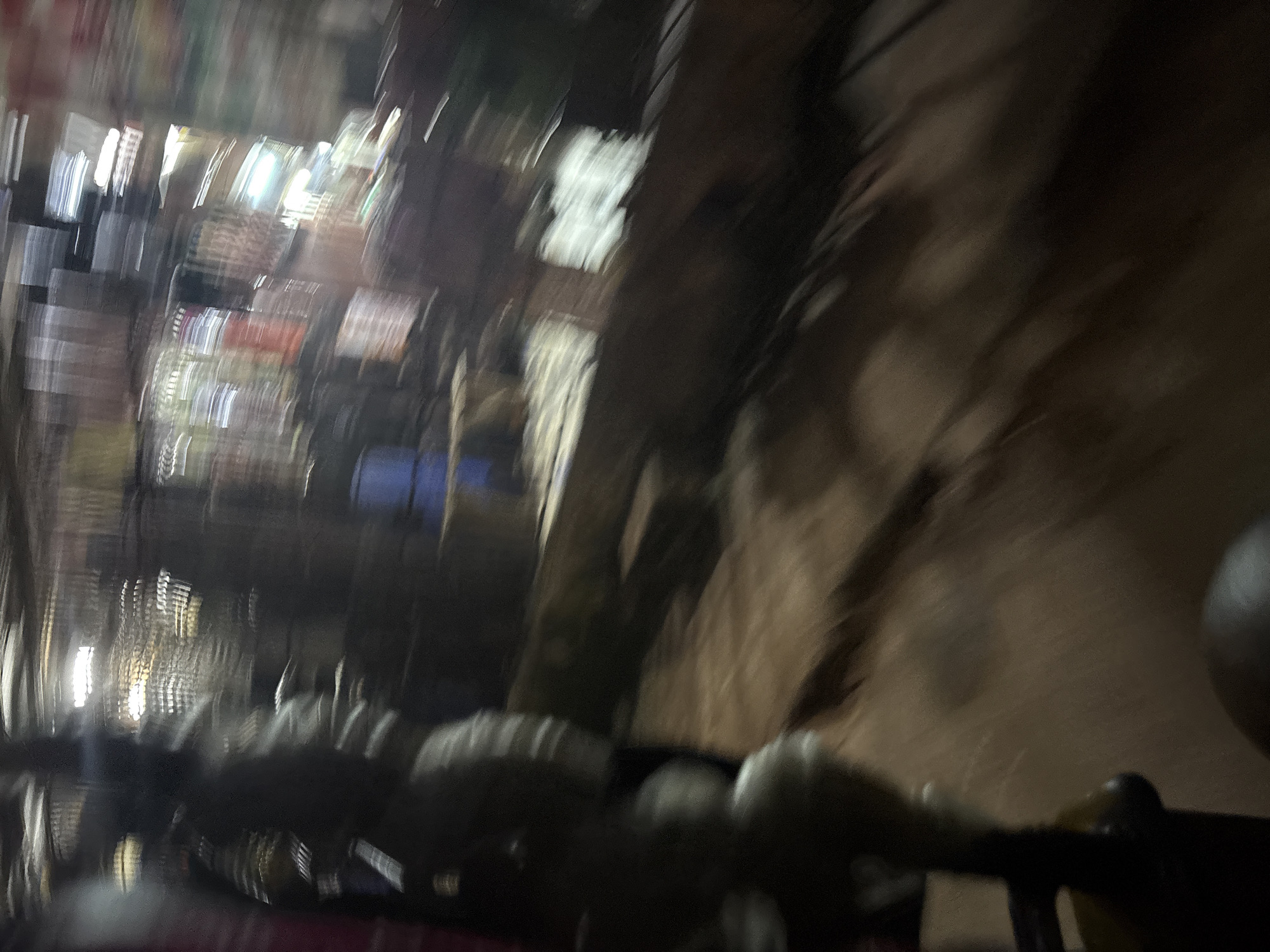 The image depicts a blurred nighttime scene that appears to be an outdoor market or street vendor area in Onitsha, Nigeria. A figure, possibly a person or a stall, is visible in the foreground, with dark clothing and indistinct features due to motion blur. Behind this figure, the background shows a series of illuminated stalls or shops, characterized by the presence of bright lights, some of which appear to be fluorescent tubes. Various rectangular shapes, likely goods or displays, are discernible within the stalls, with colors like red and white occasionally peeking through the blur. The ground appears to be rough and uneven, possibly dirt or gravel, reflecting the ambient light in streaks due to the motion blur. The overall impression is one of a bustling, active environment captured during a moment of movement, typical of a busy market at night in a Nigerian city.