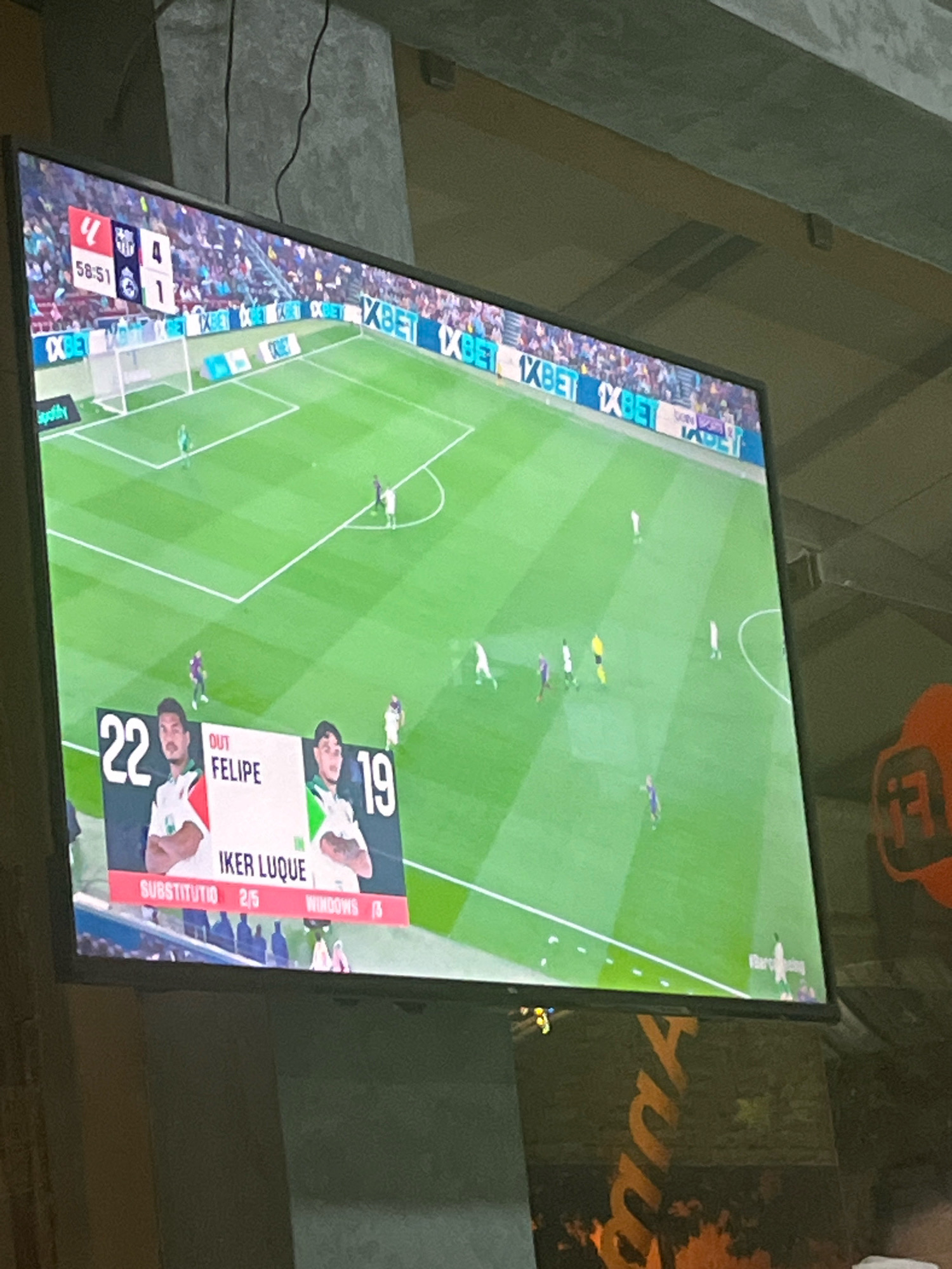 The image is a photograph of a television screen displaying a football match. The score is visible at the top of the screen: 4-1. A portion of the football field with players is visible. A graphic overlay on the bottom of the screen shows player information. One player is labeled "OUT FELIPE" and another is labeled "IKER LUQUE" with the number "19". There is also information indicating "SUBSTITUTIO 2/5 WINDOWS". The television is positioned in a room with a dimly lit, textured ceiling and what appears to be a concrete pillar. The photograph is taken from an angle, suggesting the viewer is sitting and looking up at the screen. There are no visible elements in the image that specifically place it in Azilal, Morocco; the location is inferred from the prompt.