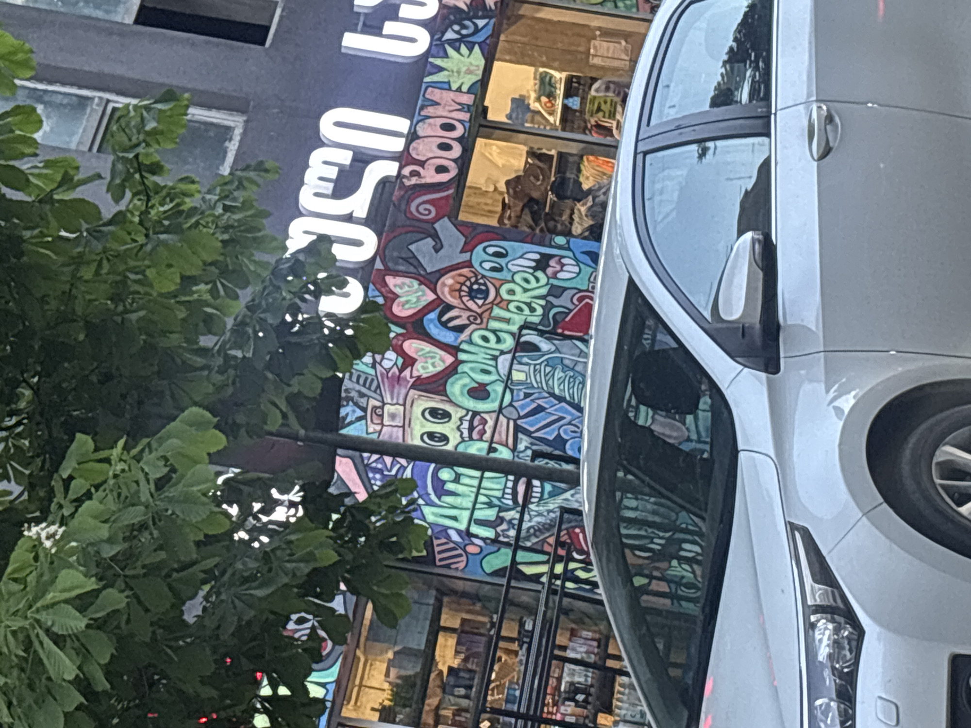 The image presents an urban street scene in Tbilisi, Georgia, dominated by a vibrant building facade and a modern vehicle, viewed from a slightly oblique angle.
On the left, lush green foliage from a tree partially obscures the upper portion of a building with a dark-colored facade. Prominently displayed on the building are large, glowing white letters in Georgian script. While partially covered, fragments of words such as "ᲗᲣᲚ" are discernible. Beneath this signage, the building's lower facade is covered in a dynamic and colorful mural or graffiti. This artwork features a variety of cartoon-like characters, hearts, eyes, and abstract shapes rendered in bright hues of green, blue, pink, and red. Within the mural, the word "BOOM" is clearly visible, oriented vertically, and another section appears to spell "somewhere" or "some where" horizontally. Behind this artistic display, shop windows reflect the interior, revealing shelves stocked with items, suggesting a retail business.
To the right, a light-colored, possibly white or silver, vehicle, resembling an SUV or minivan, is prominently featured. It is positioned with its side facing the viewer, partially overlapping the colorful building facade. Visible details of the car include its side mirror, a chrome door handle, and part of its front headlight assembly. Reflections in the car's windows show elements of the surrounding environment, including the trees and parts of the mural.
The scene appears to be set during dusk or evening, indicated by the glowing signage. No individuals are visible, and the image captures a static moment of the streetscape. The strong visual elements of the Georgian script and the intricate graffiti mural provide distinct location cues.
