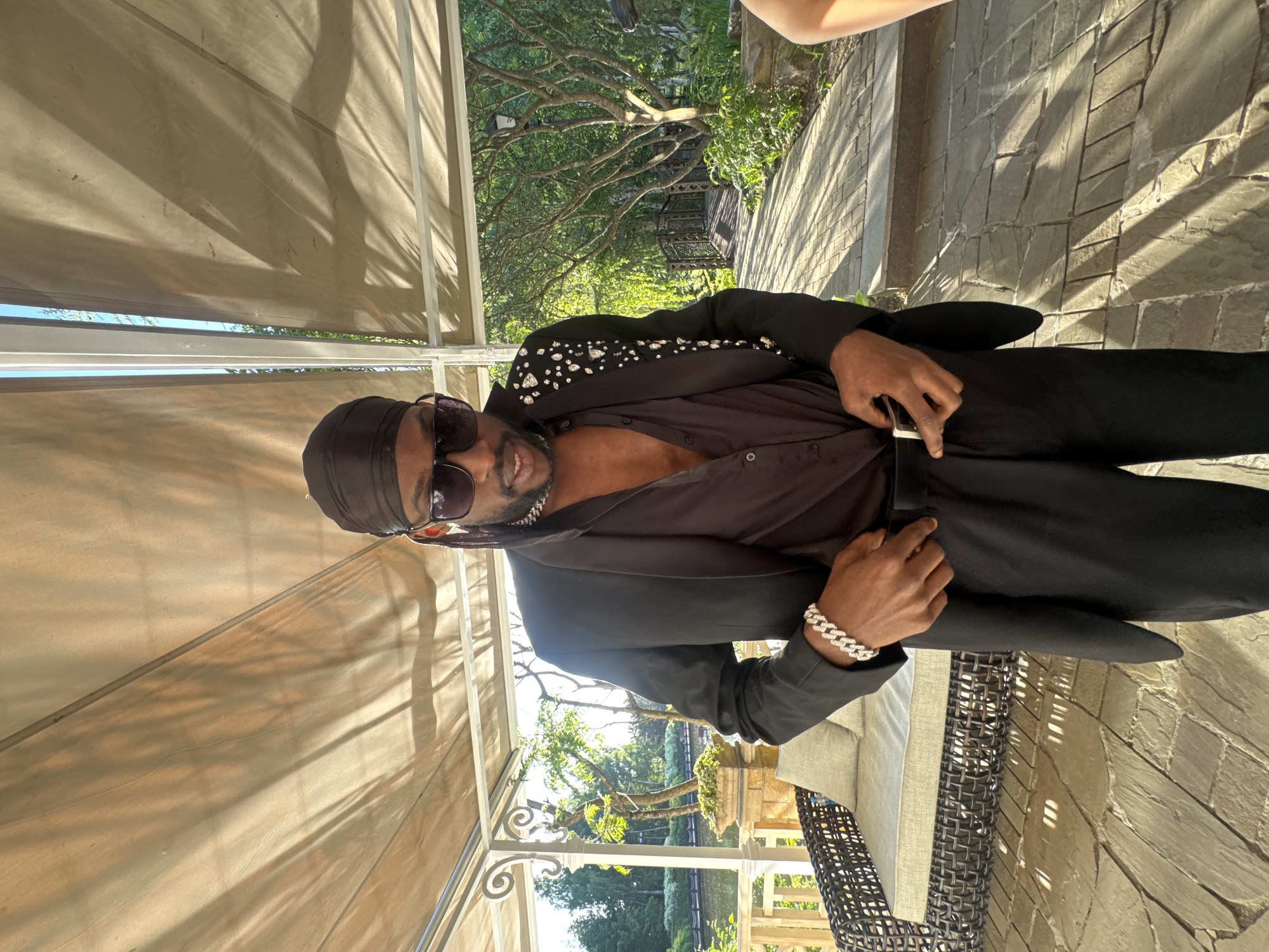 The image depicts a single male individual standing outdoors under a covered patio structure in Zhavoronki, Russia. The man is positioned centrally, looking downwards, and is actively adjusting his black trousers or belt with both hands at the waistline. He is wearing a black durag or headwrap, dark sunglasses, a silver chain necklace, and a partially unbuttoned black collared shirt beneath a black blazer. The left lapel and shoulder of the blazer are embellished with numerous reflective elements, resembling rhinestones or studs. A silver-toned Cuban link bracelet is visible on his left wrist.
The immediate ground surface is paved with irregular flagstones. Overhead, a light-colored fabric canopy or awning, supported by white metal framing, casts distinct shadows. To the left, a wicker outdoor lounge chair with light grey cushions is situated, adjacent to decorative white metal railings that form part of the patio structure. Beyond the patio on the left, a landscaped area with lush green trees, foliage, and a body of water is visible. To the right of the subject, a stone-paved pathway extends into the background, flanked by abundant greenery. Further along this path, a dark metal bridge with ornate railings is present. The scene is illuminated by natural sunlight, creating prominent shadows consistent with either morning or late afternoon.