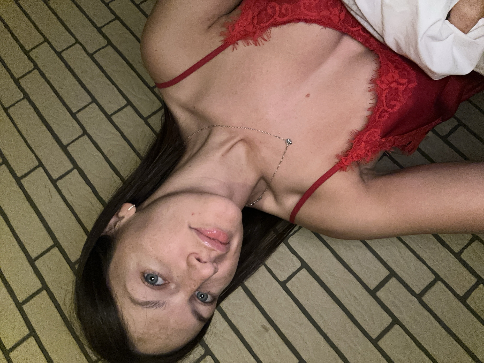 The image is a close-up selfie of a woman with long, dark hair. She is lying down and wearing a red lace camisole. She has blue eyes, freckles, and is wearing a delicate necklace. The background appears to be a textured wall with a brick pattern. The image does not contain any information about the city of Fredenbeck, Germany.