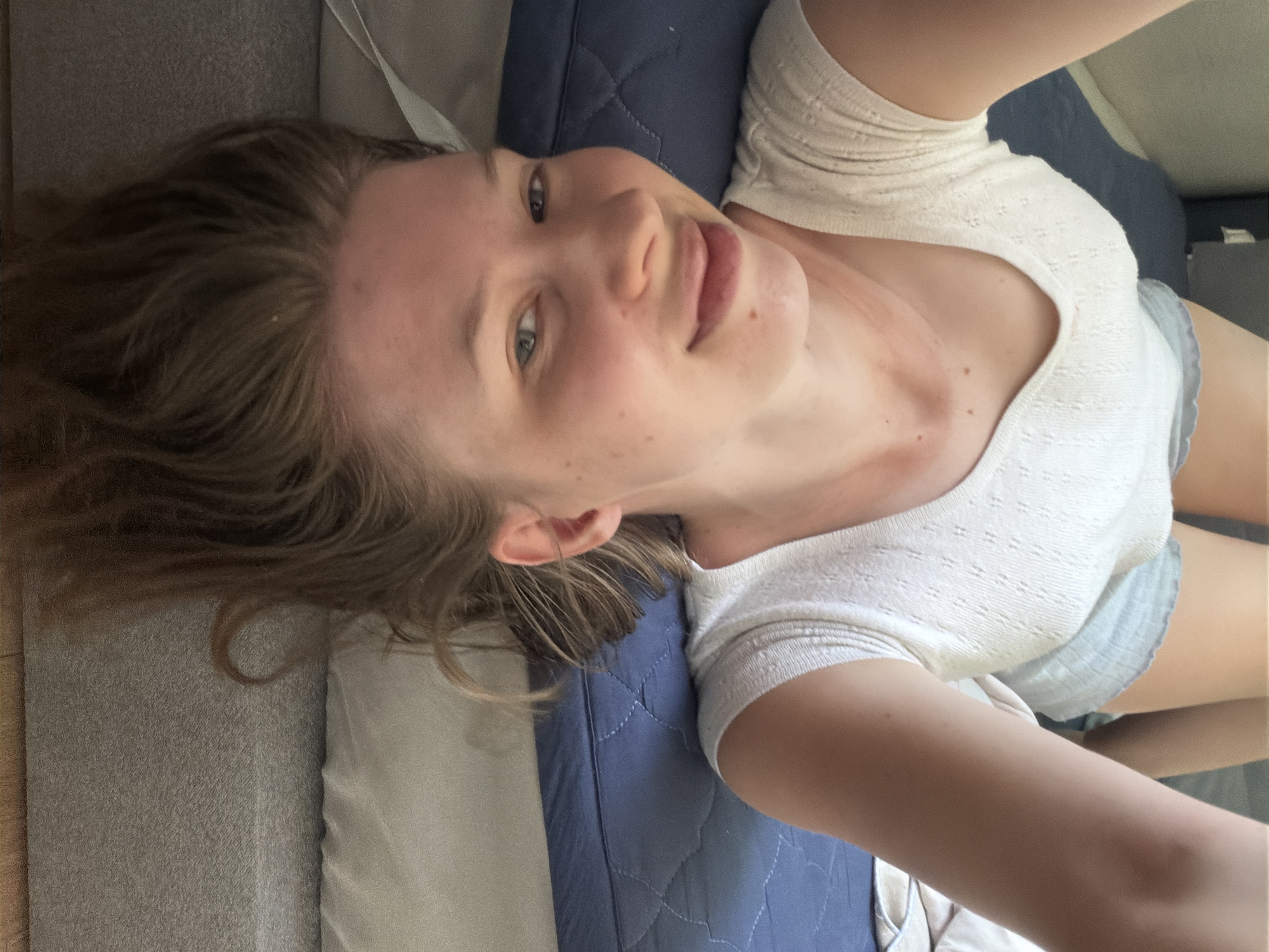 The image shows a young woman lying on her back on a bed with her head upside down. She is wearing a white top and light blue shorts. Her arms are raised above her head. The background is a bed with a blue mattress and gray bedding. The room appears to be a bedroom. The image does not provide any information about the city of Dongducheon-si, South Korea.