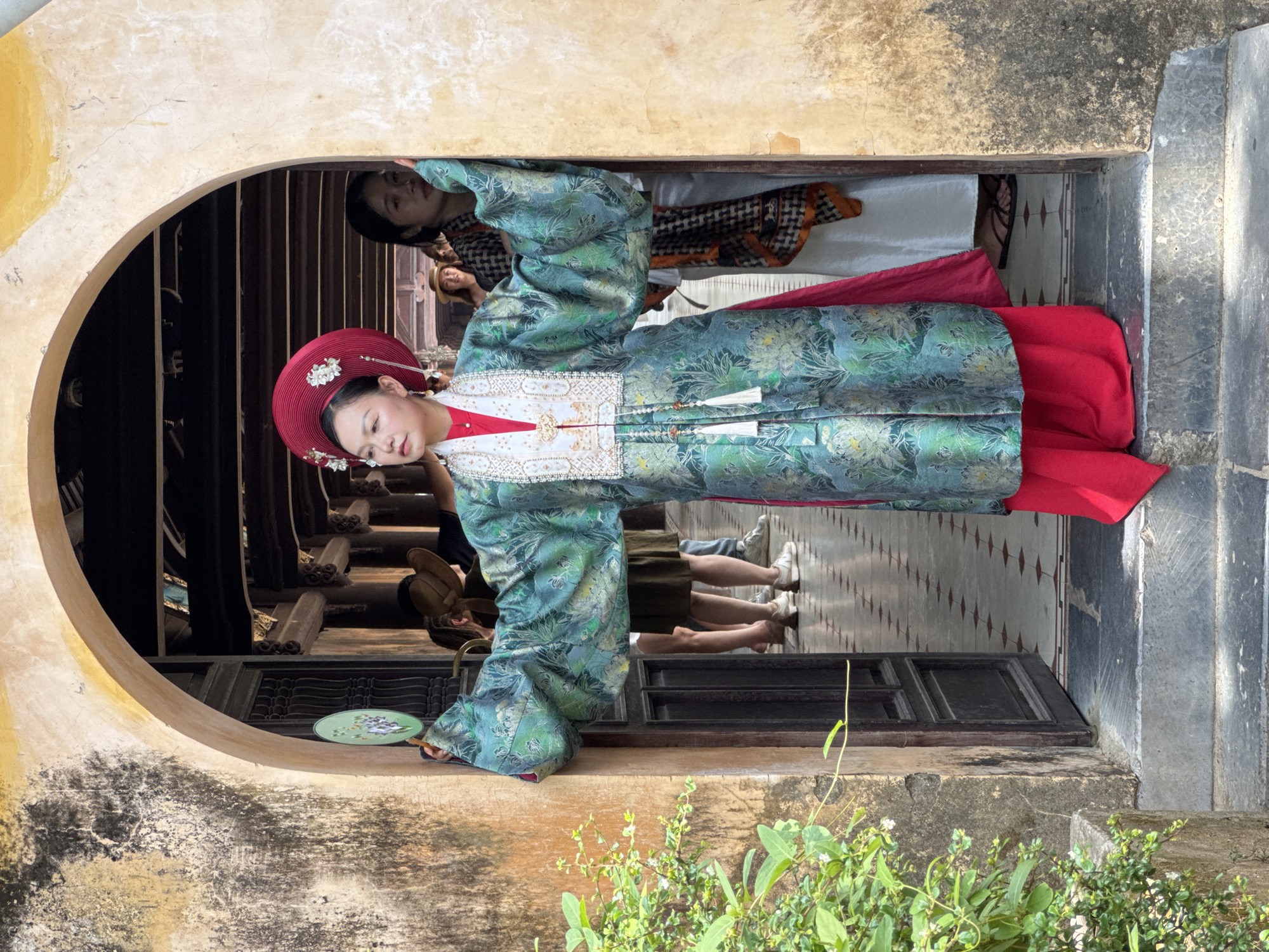 A woman in traditional Vietnamese clothing stands in a doorway. She wears a red headpiece adorned with white flowers and a long, teal robe with a floral pattern. Beneath the robe, a bright red skirt is visible. She holds a green fan in her left hand, which is partially obscuring a doorway behind her. Another woman, wearing a patterned scarf, stands to her right, partially in shadow. The doorway leads to a long hall with a tiled floor and wooden beams overhead. The doorway is framed by a stone archway with peeling yellow paint. Green foliage grows to the left of the doorway. The scene is likely set within an historical building in Phú Xuân Ward, Vietnam, given the architecture and traditional attire.