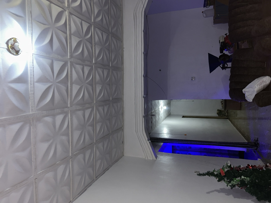 A room with textured white wall panels and a ceiling light is adjacent to another room through an arched doorway. The second room contains a brown sofa, lighting equipment, and miscellaneous items. Further down, the room extends into a blue-lit area with a Christmas tree in the corner. The spaces appear to be brightly lit, creating a sterile and modern aesthetic.