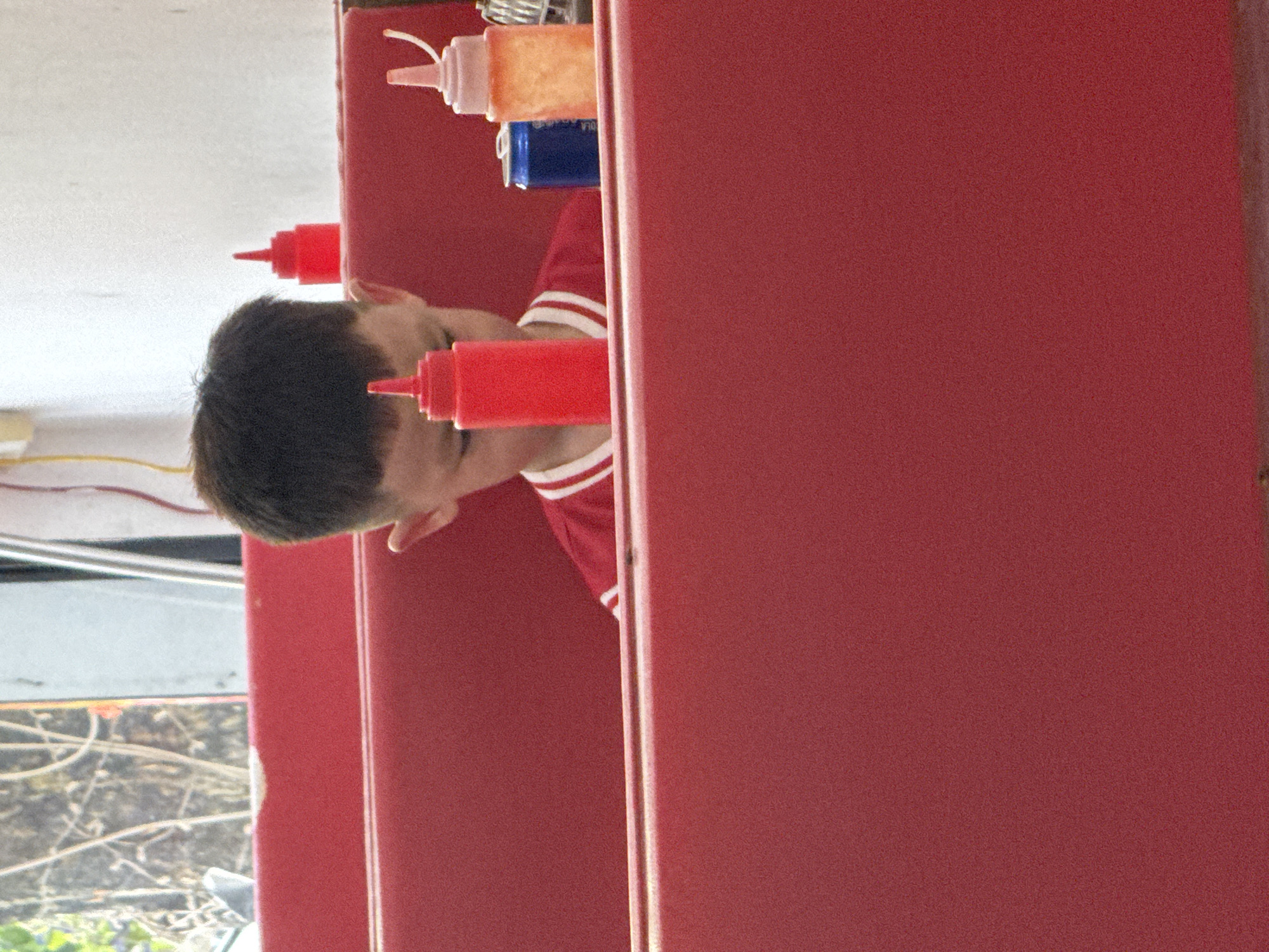 The image displays a young male seated in a red booth within a casual dining establishment located in Trung Tam Ward, Vietnam. The individual's head, featuring short dark hair, is visible above a red upholstered partition, with his face obscured as he looks downwards. He wears a red shirt with white horizontal stripes on the collar. On the red table or ledge in front of him are three condiment bottles: a prominent red squeeze bottle in the foreground, a smaller red squeeze bottle further back, and a translucent orange/pink squeeze bottle. A blue soda can rests between the prominent red bottle and the orange/pink bottle. To the left, a window provides a blurred view of outdoor foliage, primarily tree branches. To the right, a plain white wall is visible, with yellow and red electrical wires running vertically near the top. The scene is brightly illuminated, characteristic of an indoor public eatery.