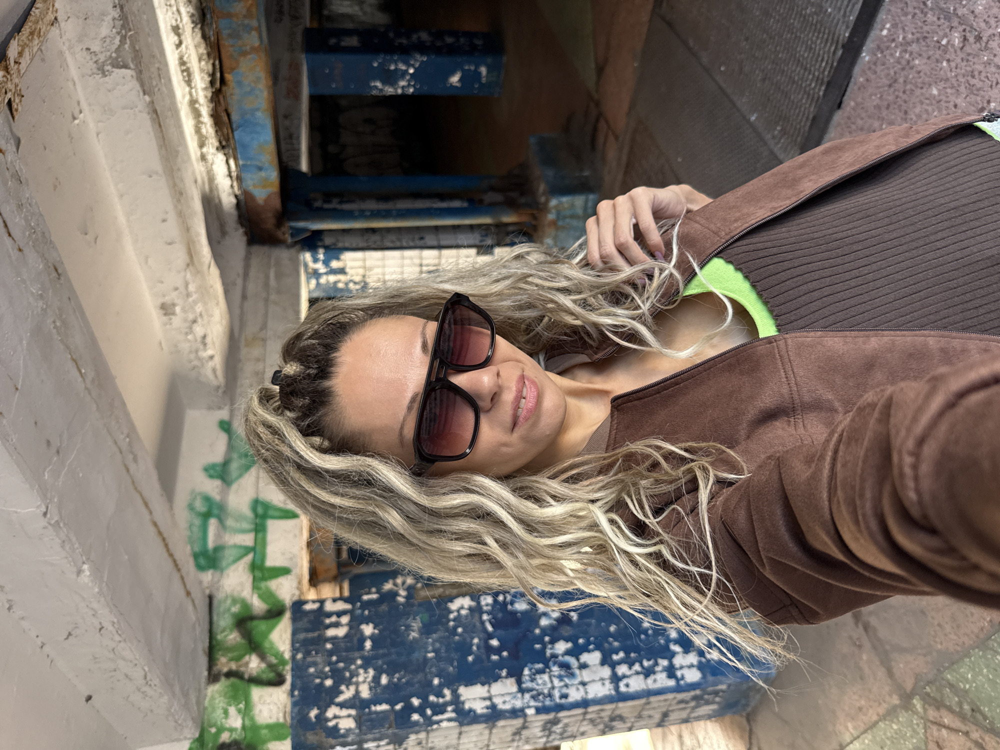 A woman with blonde, curly hair pulled into a ponytail is taking a selfie in what appears to be an urban, possibly abandoned, location. She is wearing sunglasses, a brown jacket, and a green and brown ribbed top. The background features graffiti on blue and white tiled walls, concrete structures, and rusty metal elements. The lighting suggests an outdoor or semi-outdoor setting with overhead structures. The ground appears to be paved with rough stones or concrete. The overall impression is one of a gritty, urban environment. The image does not contain any specific visual cues indicating the city of Zhukovo, Russia.