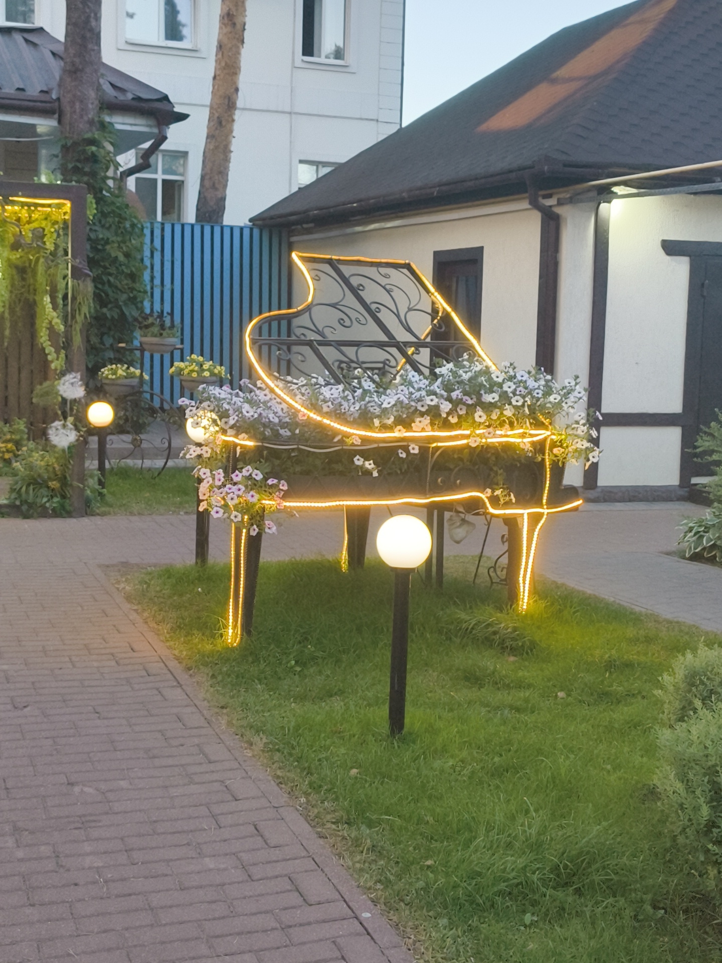 The image shows an outdoor decorative piano structure with yellow lights, filled with white and pink flowers. The piano is situated on a grassy area next to a brick pathway. In the background, there is a blue fence and buildings with dark roofs. A round, white lamp post is visible in front of the piano. The scene appears to be an exterior garden or courtyard. The location is identified as Ramenskoye, Russia.