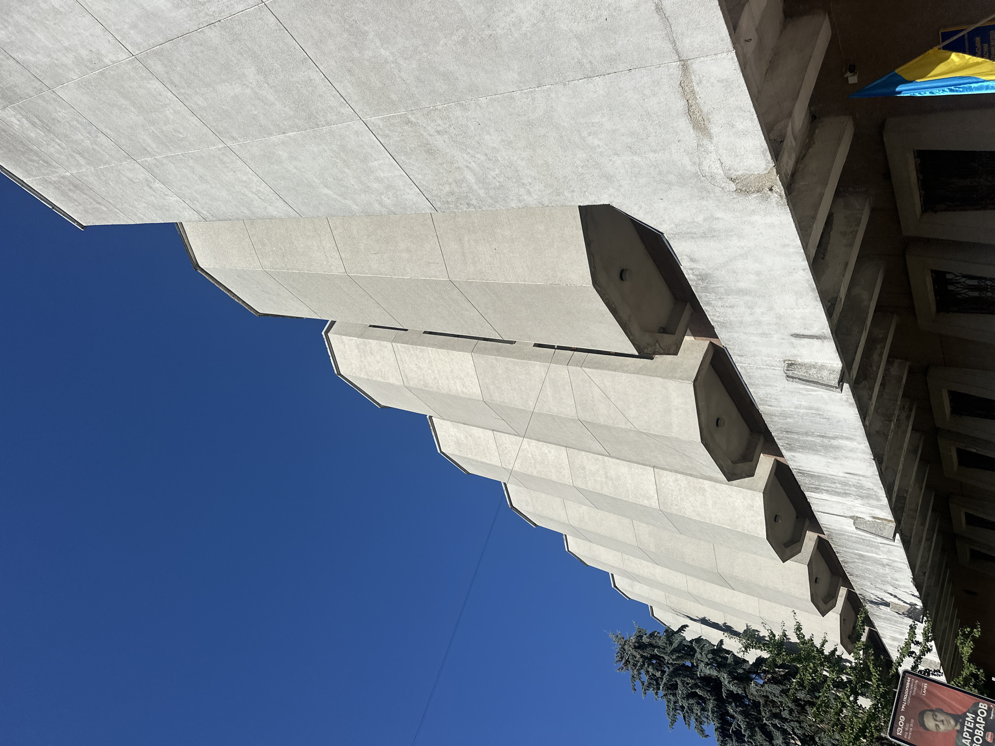 The image shows a concrete building with a distinctive geometric facade against a clear blue sky. The building's design features a series of angled panels that create a rhythmic, stepped appearance. Some of these panels extend outwards, forming recessed balconies or alcoves. Below the main facade, there is a concrete overhang supported by angled beams, with windows visible beneath it. A tall evergreen tree is positioned to the left of the building. A red poster with a portrait of a man and some text is partially visible on the left side, suggesting a public event or advertisement. On the right side, a Ukrainian flag is attached to the building, with its blue and yellow colors prominent. The architecture appears to be modern, possibly from the mid to late 20th century, and the overall scene is well-lit, indicating daylight.
The building in the image is the Ternopil Regional Philharmonic Hall, located in Ternopil, Ukraine.