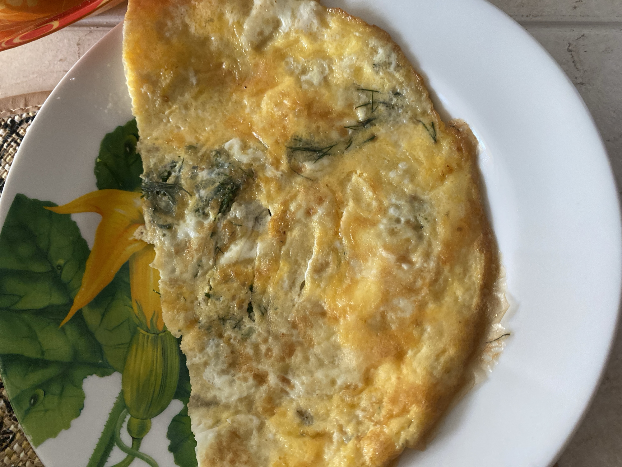A close-up, top-down shot shows a folded omelet with green herbs on a white plate with a decorative pattern of green leaves and yellow squash flowers. The omelet is golden brown with some lighter and darker spots, indicating it is cooked through. The plate has a glossy finish, and the background is a woven placemat. The scene appears to be indoors, likely in a home setting. There are no people or animals visible. The location is identified as Lida, Belarus.
