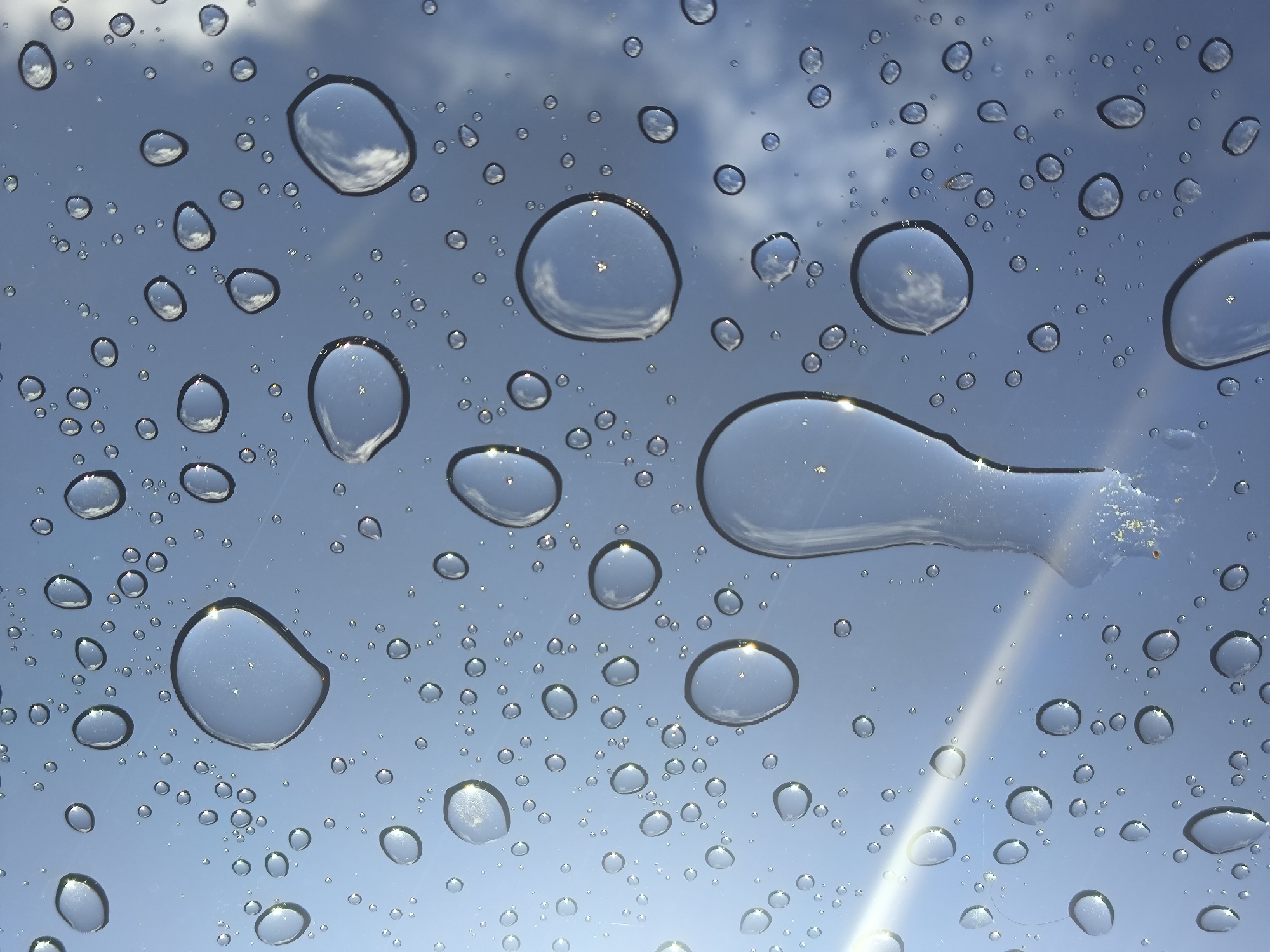 The image shows a close-up view of water droplets on a glass surface, likely a window or a car windshield. The droplets vary in size and shape, with some appearing spherical and others more irregular. Each droplet acts as a lens, distorting and magnifying the reflection of the sky behind it. The sky is a pale blue with scattered white clouds, suggesting an overcast or partly cloudy day. Sunlight refracts through some of the larger droplets, creating bright highlights and streaks of light. The overall impression is one of texture and light play, characteristic of a rainy or humid environment. There are no people, animals, or man-made objects other than the glass surface itself visible in the image. There is no information in the image that would indicate its location in Kaliningrad, Russia, or any other specific city.