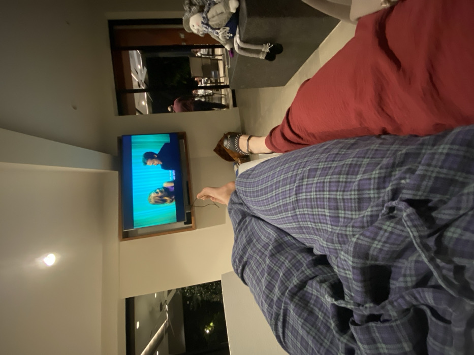 A person wearing plaid pajama pants is lounging in front of a television, which displays a scene from a show or movie. Another person wearing a red garment sits beside them. A doll sits on a surface near the television. Through a window, greenery and exterior lights are visible, while the interior features a modern design.
