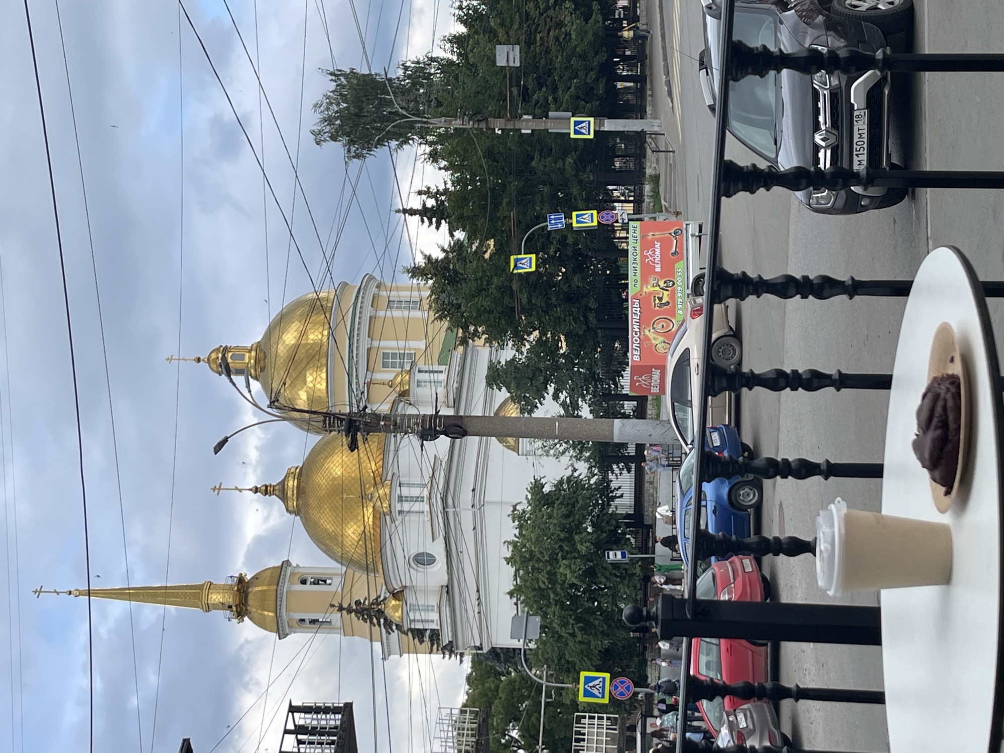 The image captures a street scene in Izhevsk, Russia, with a prominent golden-domed church in the background. The church features a tall, slender spire topped with a cross and multiple rounded domes, also adorned with crosses. The building itself is white with yellow accents. In the foreground, a black metal railing separates the viewer from a street with several cars, including a red hatchback, a blue hatchback, and a dark grey Renault Duster. A white cup and a chocolate pastry are placed on a white table in the immediate foreground, suggesting a cafe setting. Pedestrians are visible on the sidewalk near the church and crossing the street. Road signs, including pedestrian crossing signs, are present. The sky is overcast with clouds. A banner advertising bicycles is visible on the side of a vehicle in the mid-ground. The license plate of the Renault Duster reads "M 150 MT 18".