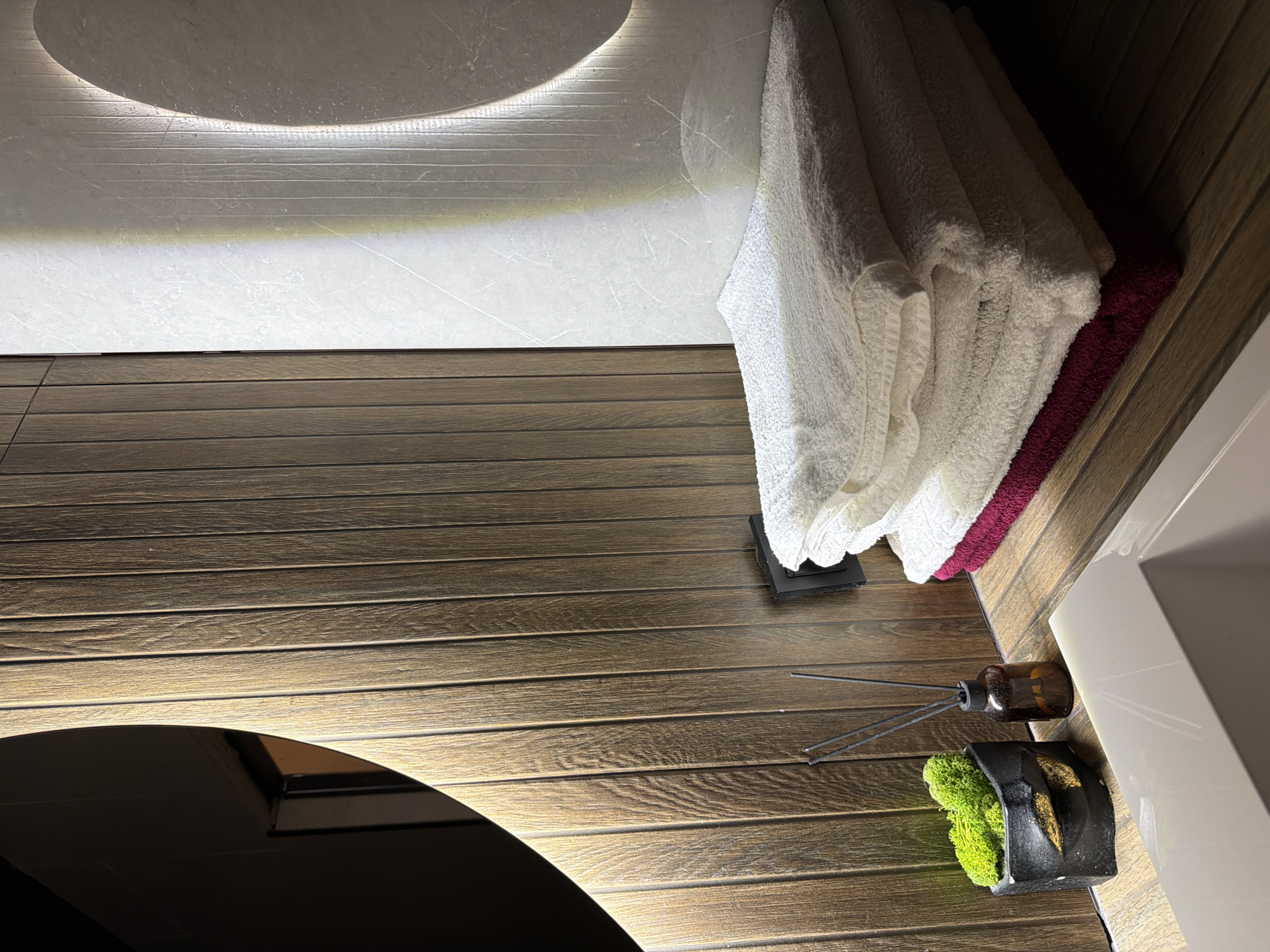 This image depicts a close-up view of a bathroom vanity area with a modern aesthetic. The surface appears to be made of horizontally slatted wood. On the left side, a black planter with a gold-colored accent holds vibrant green moss. Next to it is a small amber glass bottle with dark diffuser reeds, suggesting an air freshener. In the background, a wall features vertical wooden paneling and a section of light-colored textured material with vertical grooves. A large, circular mirror with integrated lighting is partially visible on the left, reflecting a dark interior space. To the right, a stack of folded towels is prominently displayed. The top two towels are white and appear to be made of terrycloth. Beneath them is a dark red towel, and possibly another towel underneath that is mostly obscured. A light-colored sink basin is visible in the foreground. The lighting in the scene is warm and casts distinct shadows, highlighting the textures of the materials. There are no people present, and no discernible actions or interactions are taking place. The image is a still life composition focused on the decorative and functional elements of a bathroom. This scene is not identifiable as being located in the city of Семилуки, Russia.
