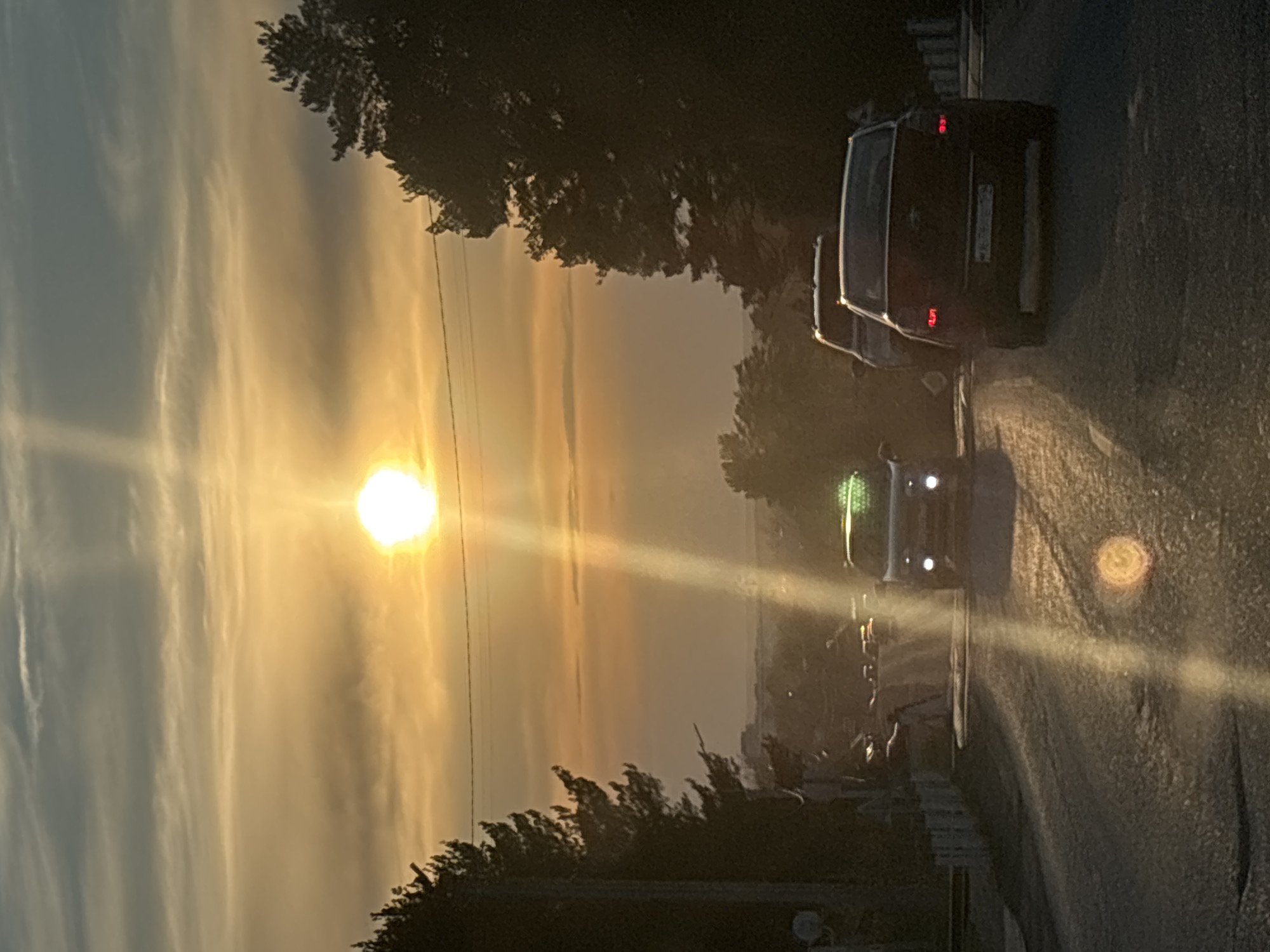 The image captures a vibrant sunset over what appears to be a roadway in the city of Chaikovsky, Russia. The sun dominates the sky, casting a golden glow and creating strong lens flare effects. Wispy clouds are visible near the sun.
A line of trees is silhouetted against the bright sky, lining the road on both sides. Several cars are visible on the road, with their headlights adding to the overall light. One car is closer to the viewer, parked on the right side of the road. Other buildings are visible in the distance. The photo is taken from a low angle.