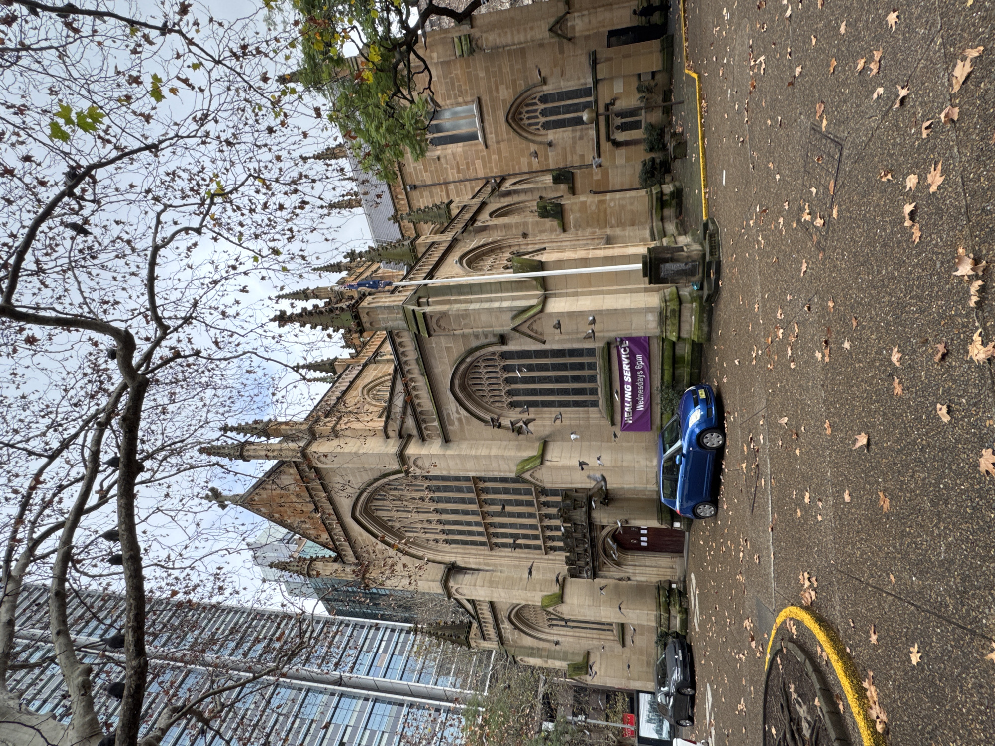 In Sydney, Australia, a grand Gothic Revival style cathedral, St. Andrew's Cathedral, is captured in a low-angle, full shot. The sandstone structure is adorned with intricate architectural details, including spires, arched windows, and a prominent cross atop its facade. The cathedral is situated amidst an urban landscape, with a modern high-rise building visible in the background, creating a striking contrast between old and new architecture.
In front of the cathedral, a paved courtyard is scattered with fallen leaves, suggesting an autumn or winter setting. Two cars are parked on the paving: a dark grey sedan to the left and a bright blue hatchback to the right. A purple banner hangs on the cathedral wall, reading "HEALING SERVICE Wednesdays 6pm". The sky above is overcast and cloudy, contributing to the muted color palette of the scene. Several pigeons are perched on tree branches in the foreground and can be seen in flight around the cathedral, adding a dynamic element to the composition. A person in dark clothing is visible standing on the far right side of the image, near a lamppost. The foreground also features a circular planter with exposed roots and a yellow painted curb.