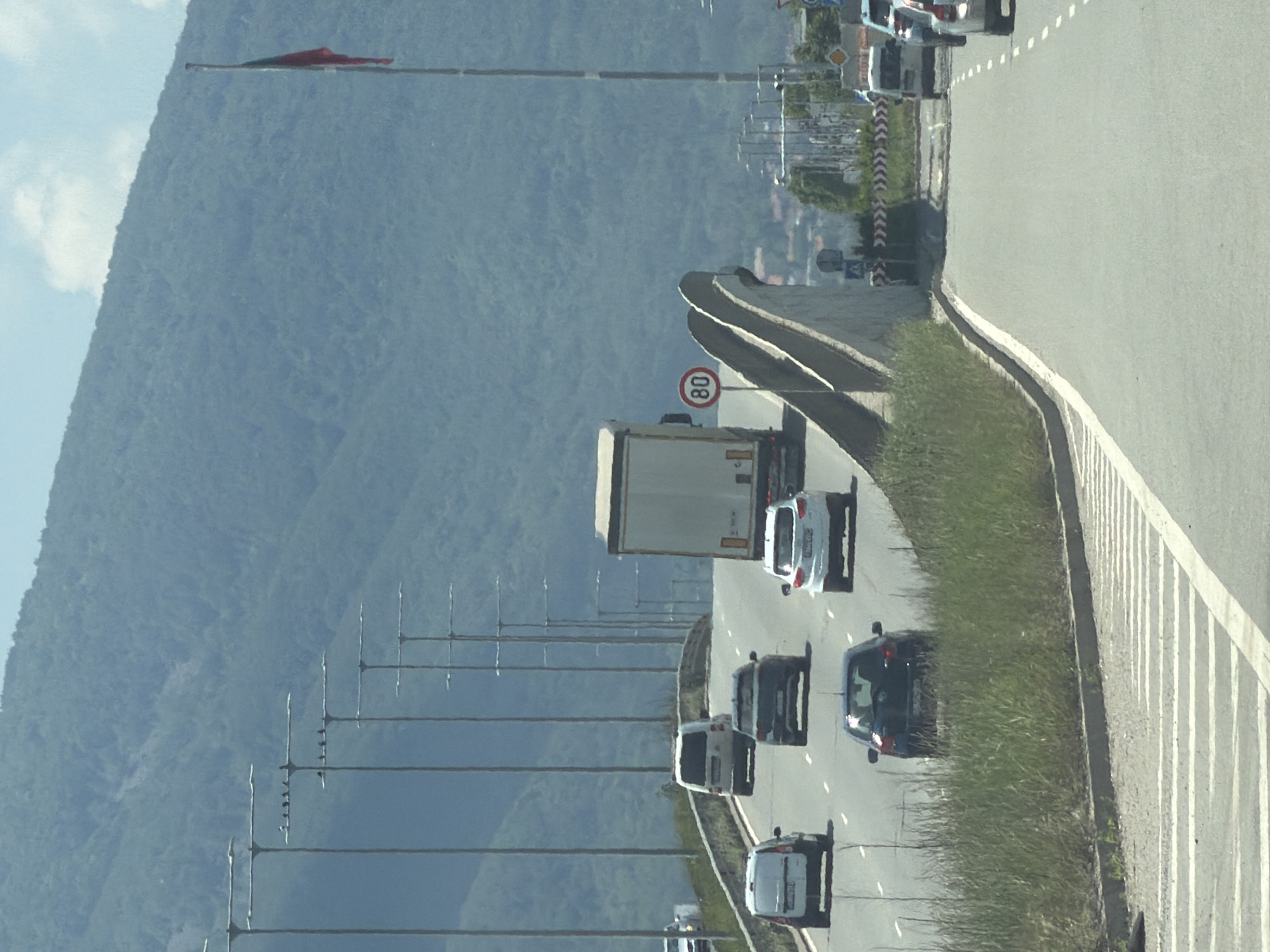 The image captures a highway overpass on a sunny day, likely in or near Sofia, Bulgaria, given the presence of a Bulgarian flag flying from a tall flagpole on the right side of the frame. The highway is paved with asphalt and marked with white lane dividers. Several vehicles are visible on the road, including a white truck in the center, a dark-colored sedan, two white vans, and a white SUV, all traveling in the same direction. A speed limit sign indicating 80 is visible on the side of the road. In the background, a lush green mountain slope dominates the landscape. Numerous tall streetlights are arranged in a line along the elevated section of the highway. The overall scene depicts a typical traffic scene on a major road with natural scenery in the backdrop.