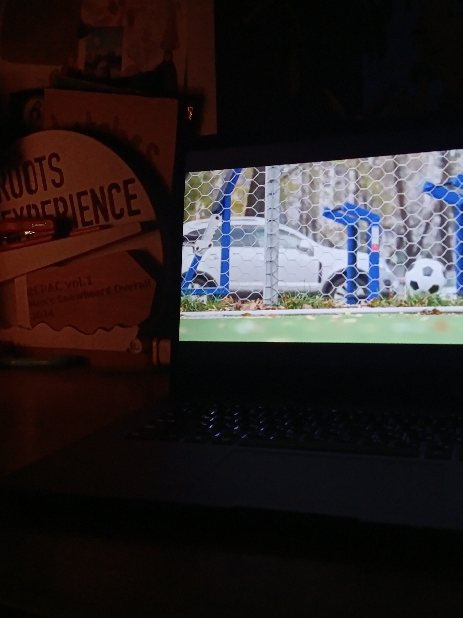 A dimly lit room features a laptop with a soccer scene displayed on the screen: a soccer ball positioned near a net, with a car visible in the background. The laptop sits on a desk. To the left of the laptop, there is a snowboard review guide.