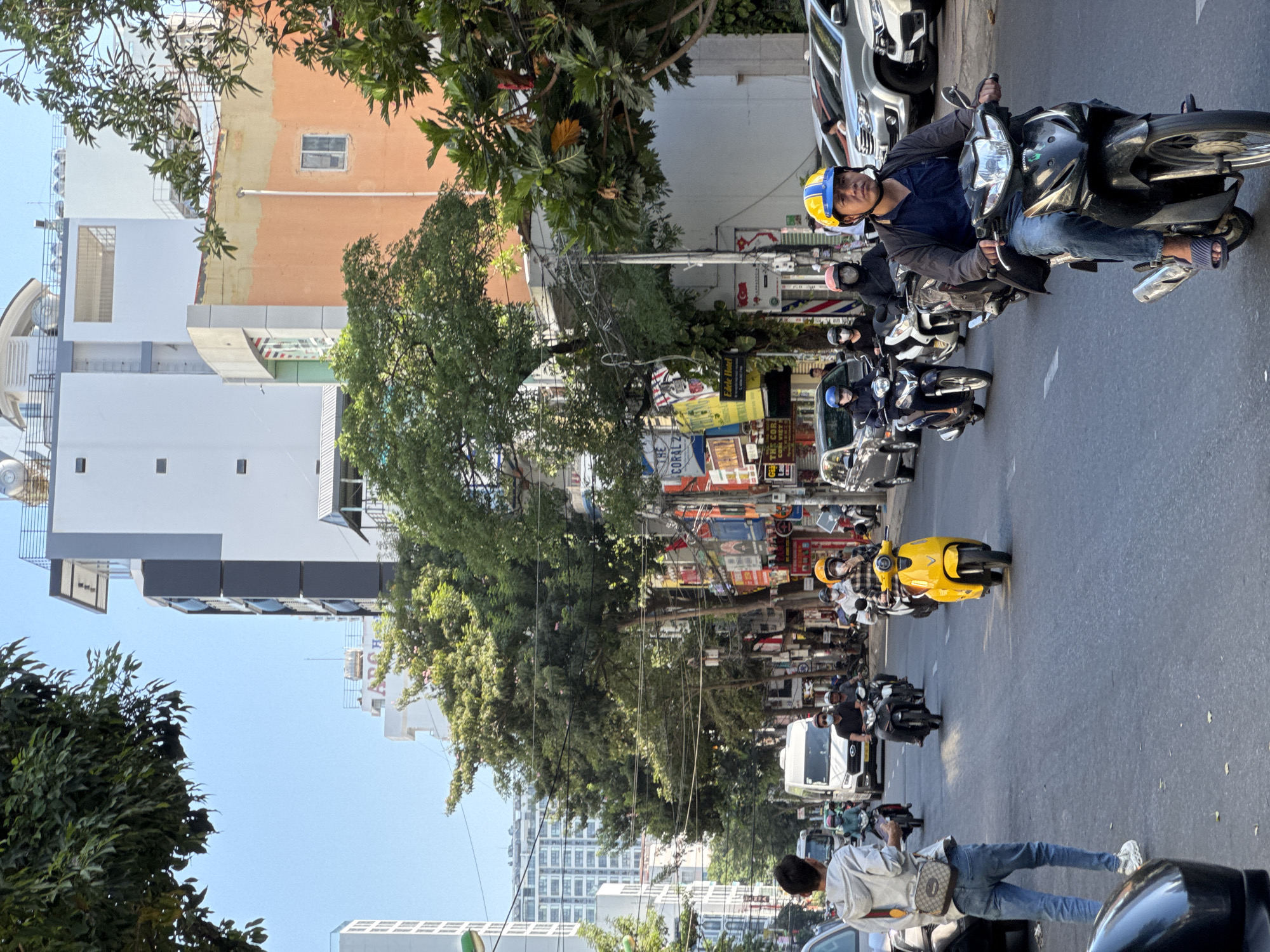 The image depicts a street scene in Nha Trang, Vietnam, with a clear blue sky overhead. The street is bustling with traffic, primarily motorbikes. In the foreground, a man wearing a blue shirt, jeans, and a yellow and blue helmet rides a black motorbike. Behind him, several other motorbikes are visible, some with riders wearing helmets. A yellow scooter is also seen in the middle ground. On the side of the street, there are various buildings, including multi-story structures with commercial signage. Trees line the street, providing shade. Cars are parked along the right side of the road. The overall impression is of a typical urban environment in Vietnam during the daytime.
