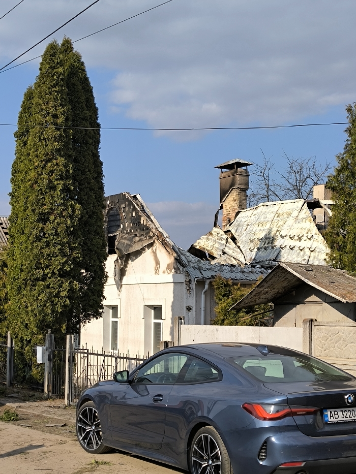 The image captures a residential street scene in the city of Vinnytsia, Ukraine, under a clear blue sky with scattered white clouds. In the foreground, a dark blue two-door BMW coupe, bearing Ukrainian license plate AB 3220 HE, is parked on an unpaved dirt shoulder, facing left with its rear visible. The vehicle appears undamaged. In the background, two residential structures exhibit significant damage. The prominent light-colored house on the left, displaying the number "57" on its facade, has a severely damaged and partially collapsed roof, with charred timbers and blackened roof materials. Smoke staining is evident on the upper exterior walls. An adjacent structure to the right also shows roof damage, with crumpled and discolored sheeting, and a brick chimney blackened by soot. A tall, slender green coniferous tree stands on the left, partially obscuring the damaged house. Dark electrical or communication lines stretch horizontally across the upper portion of the sky. No individuals are visible.