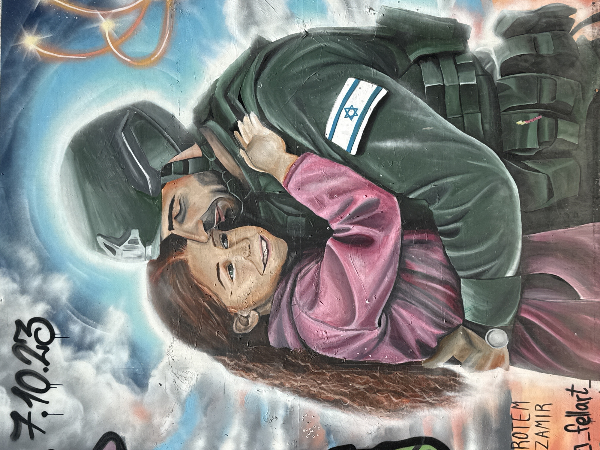 This media file displays a vibrant mural painted in Tel-Aviv, Israel, featuring a soldier embracing and protecting a child. The central figures are a male soldier and a young female child.
The soldier is depicted in a dark green military uniform, wearing a tactical helmet and body armor. An Israeli flag patch is clearly visible on his right arm. He has his arm around the child, holding her close to him in a protective gesture. The child, with long, wavy brown hair, wears a pink long-sleeved top and is looking up with a wide, radiant smile, her green eyes prominent.
The background of the mural is a bright, cloudy blue sky. In the upper left corner, several glowing orange, ring-like streaks are visible, possibly symbolizing rockets, missiles, or defensive interceptor trails against the sky.
Prominently inscribed in black on the left side of the mural is the date "7.10.23," a significant date in Israeli history. Towards the bottom right, vertical text includes the names "ROTEM ZAMIR" and "_Fellart_", likely identifying artists or individuals honored by the artwork.
The mural conveys a strong sense of protection, innocence, and resilience, with the smiling child juxtaposed against the soldier's vigilant presence and the implied threat in the sky. The art style is illustrative with visible brushstrokes and spray paint textures.