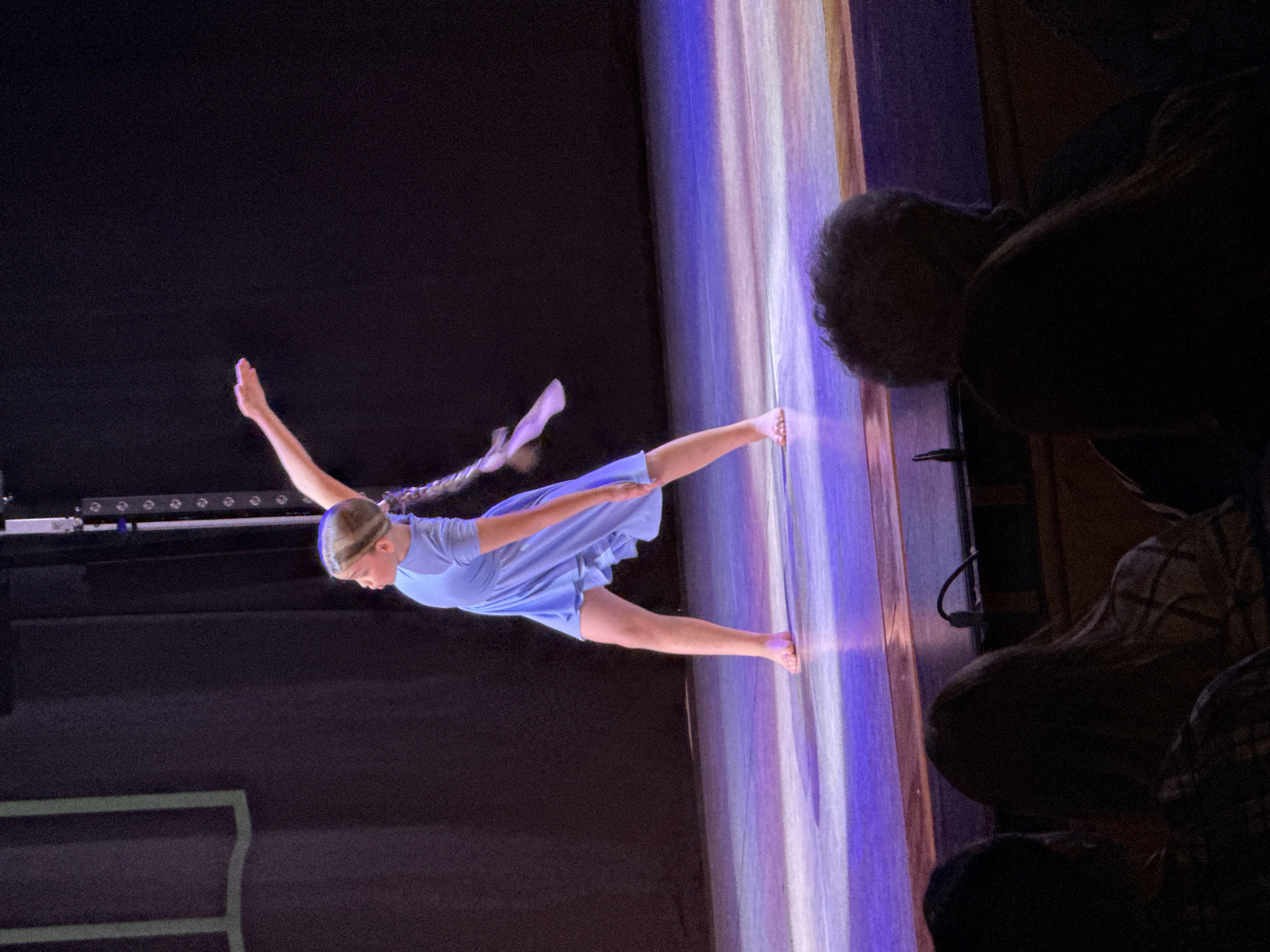 A young female dancer, wearing a blue dress, is performing on stage. She is suspended in a mid-air pose with her right arm extended upwards and right leg outstretched. The stage backdrop has vertical stripes of white, light blue and violet light. Two spectators are in the foreground in shadow. The location is not indicated.