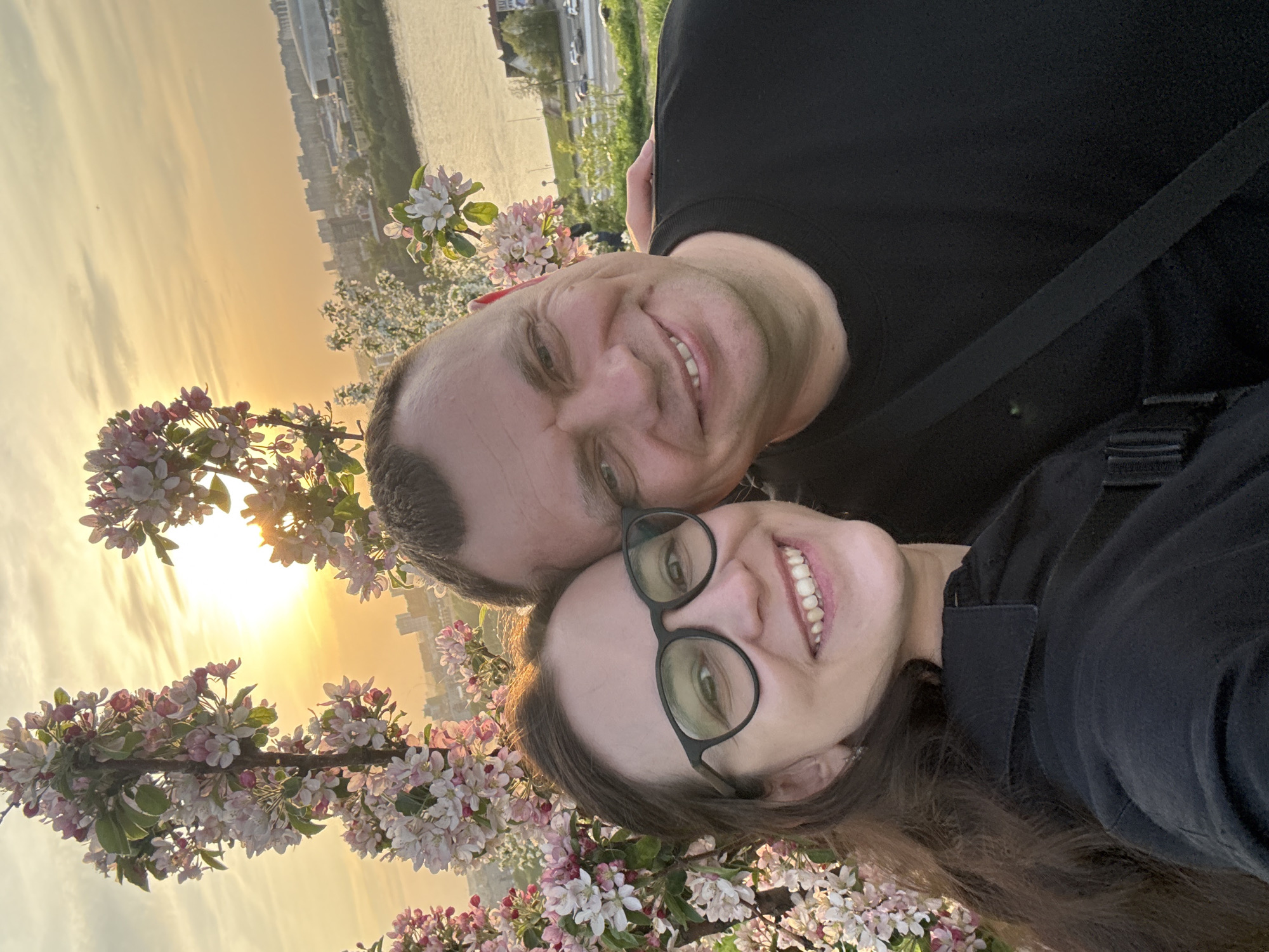 A selfie shows a man and a woman smiling at the camera in front of blossoming trees. The woman wears glasses and a black shirt. The man wears a black shirt. The sunset and the city of Nizhny Novgorod, Russia are visible in the background. Water is behind the city.