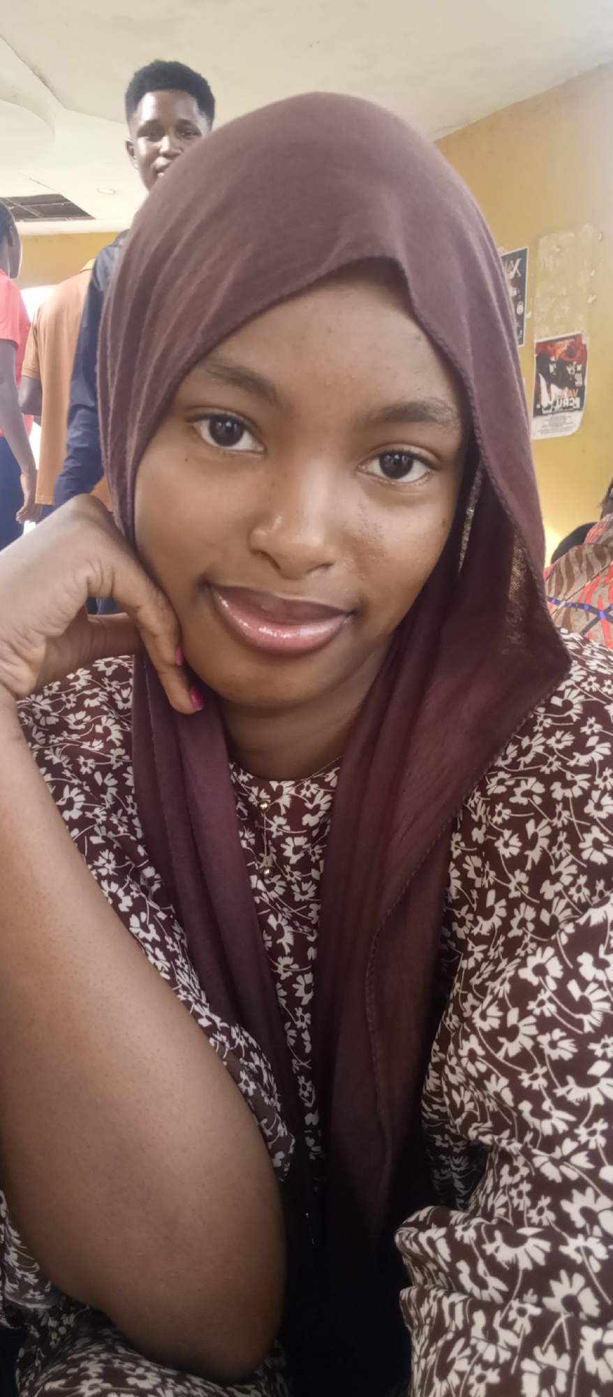 The image captures a close-up portrait of a young woman with dark skin, smiling gently at the camera. She is wearing a brown hijab that covers her hair and drapes over her shoulders, along with a brown top featuring a white floral pattern. Her right hand is propped under her chin, with a touch of pink nail polish visible on one finger. She has dark, expressive eyes and glossy lips. A delicate silver chain necklace with small golden beads can be seen around her neck.
In the background, partially visible behind her, is another young man with short dark hair, also looking towards the camera. Further to the left, parts of other individuals in red and light brown shirts are faintly visible, suggesting a gathering of people. The setting appears to be indoors, likely a casual space like a classroom or common room, given the plain walls and posters. On the wall to the right, several papers or posters are displayed; one clearly features a bold 'X', while others have unreadable text and graphics. A light fixture is visible on the ceiling in the upper left corner.
The activity depicted is a casual snapshot, with the main subject posing for the photograph. Her expression is calm and friendly. The scene suggests a relaxed social moment in an indoor environment in Keffi, Nigeria.