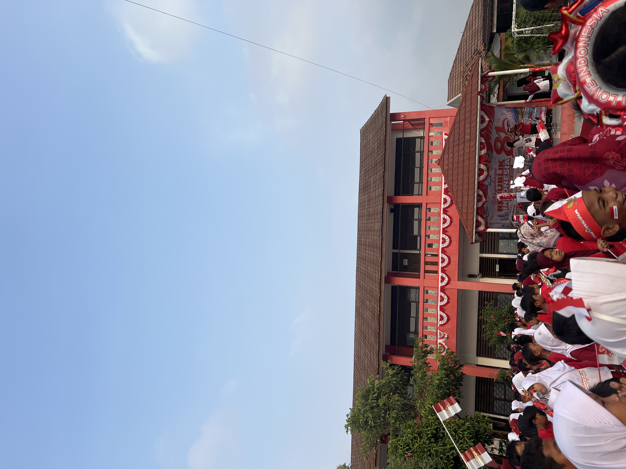 The image captures an outdoor gathering in front of a two-story red building, possibly a school, in Gelam Jaya, Indonesia. The building has a brown tiled roof and a balcony adorned with red and white bunting. A stage is set up in front of the building with a banner that reads "DIRGAHAYU REPUBLIK INDONESIA KE 81". A crowd of people, predominantly children dressed in red and white, are assembled, many wearing patriotic accessories like hats, headbands, and holding small Indonesian flags. Some children have red and white face paint. Two individuals are on the stage, one holding a microphone, suggesting a performance or speech. A group of children are in the foreground, with some looking towards the stage. The sky is clear blue with a few wispy clouds. The overall scene appears to be a festive celebration, likely for Indonesian Independence Day.